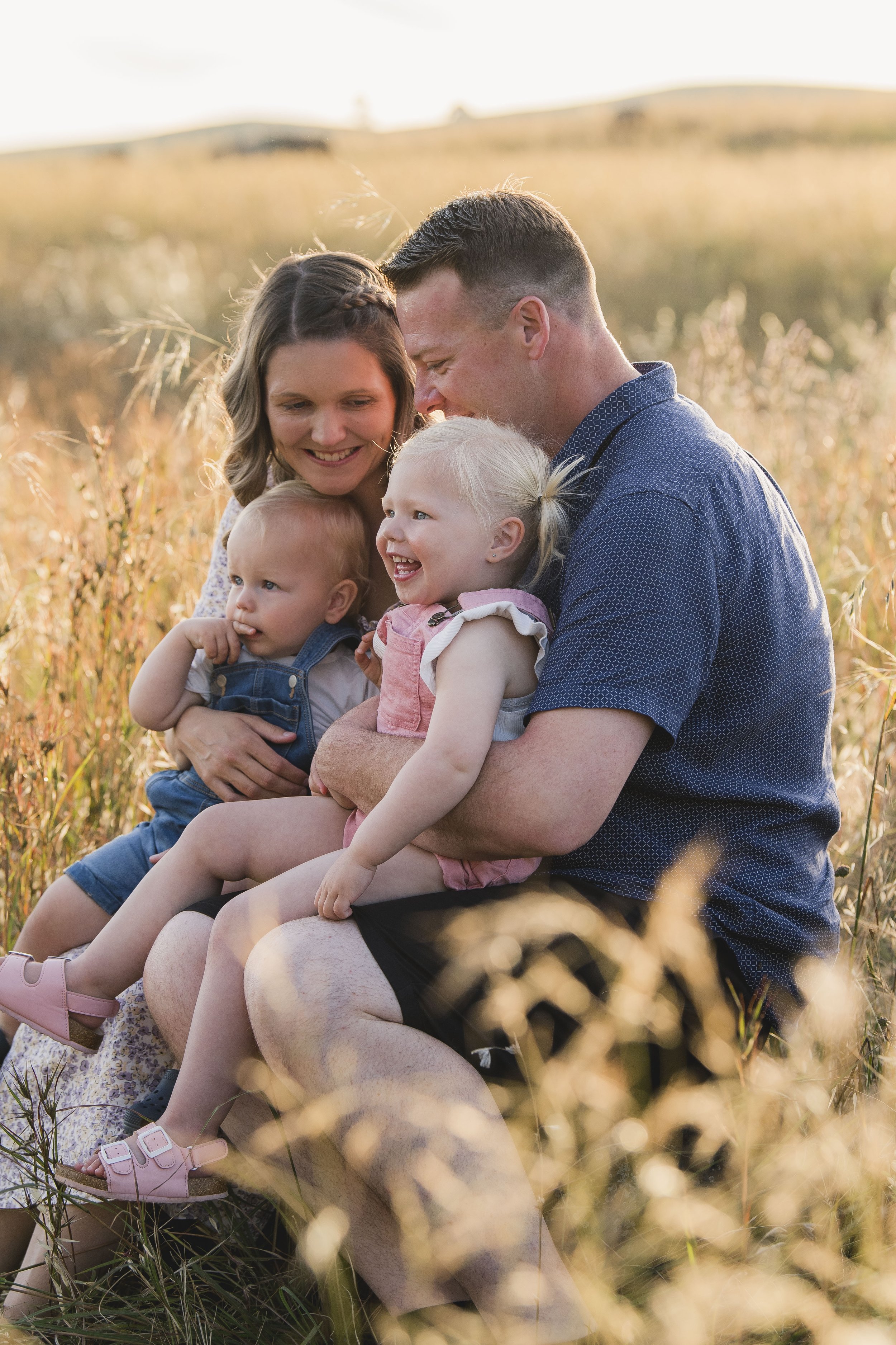 A family of four sitting in a grassy field during sunset, smiling and enjoying each other's company. The family includes a mother, father, and two young children.