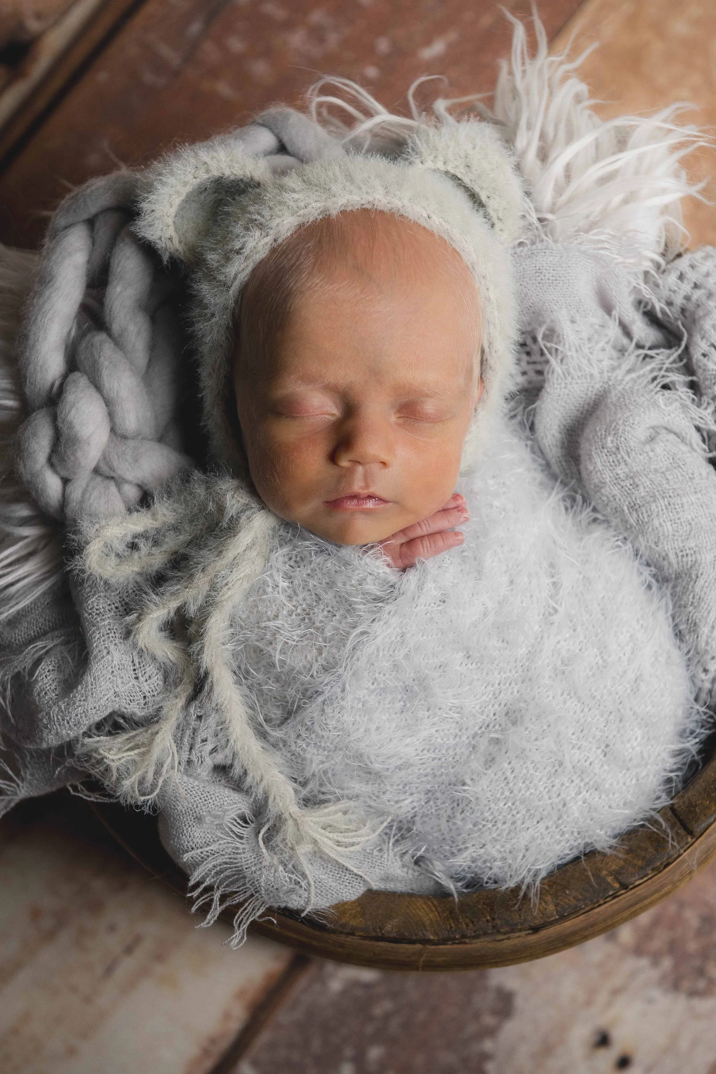 A sleeping baby with light skin and red hair, wearing a fuzzy hat with pom-poms, lying on a soft, textured blanket on a rustic wooden surface.