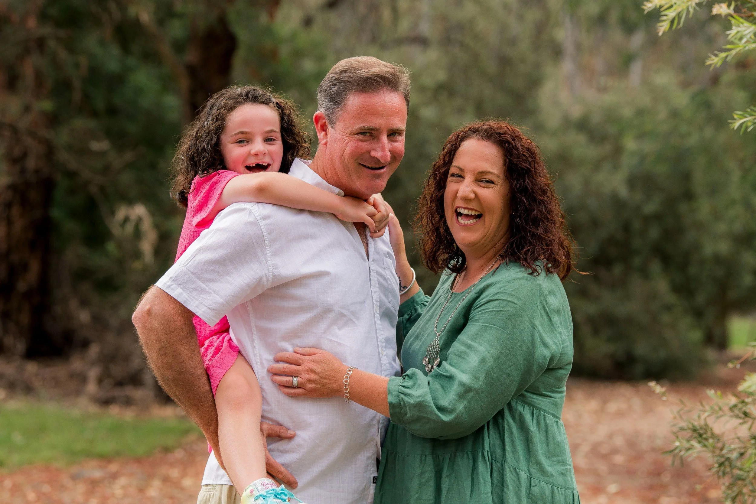 A family of three, consisting of a man, woman, and young girl, standing outdoors in a wooded area, smiling and enjoying their time together.