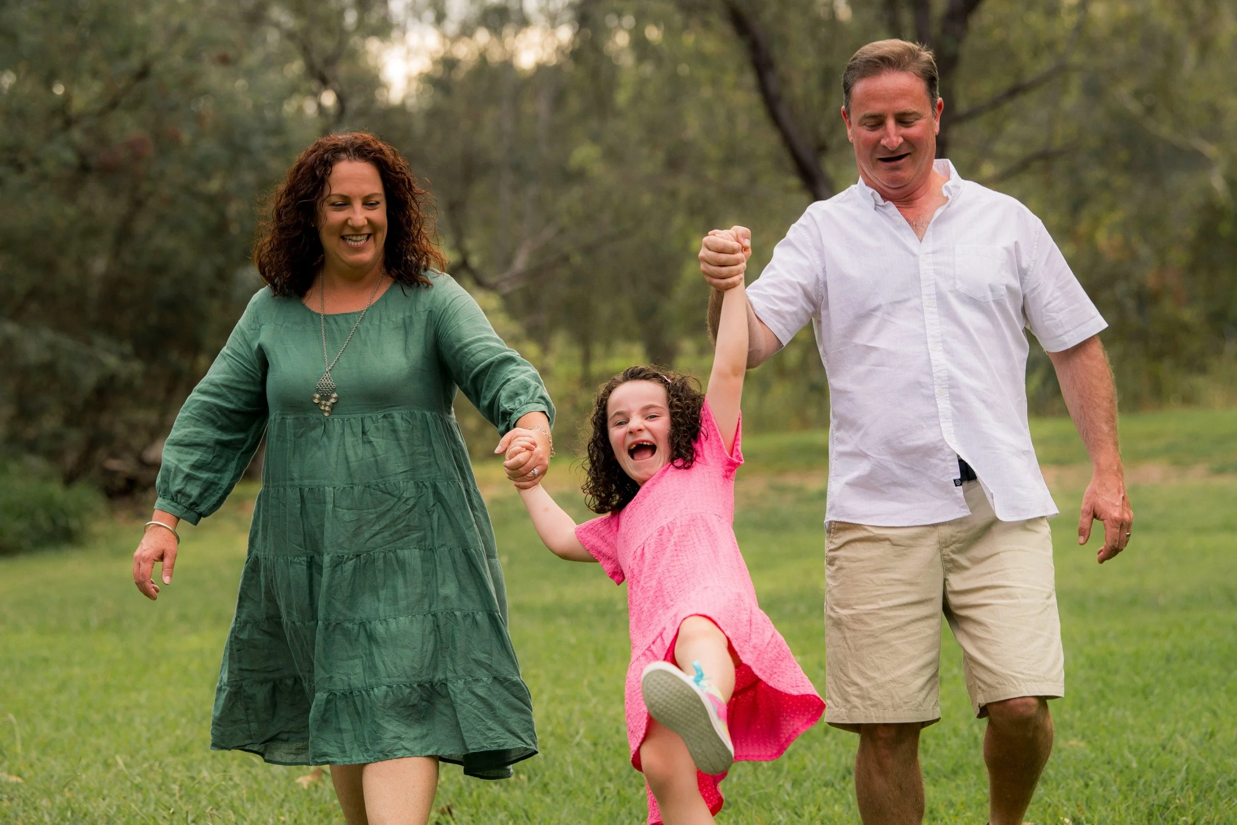 A family of three including a mother, father, and a young child, are walking happily outdoors in a park surrounded by trees. The child is smiling and holding hands with the parents.