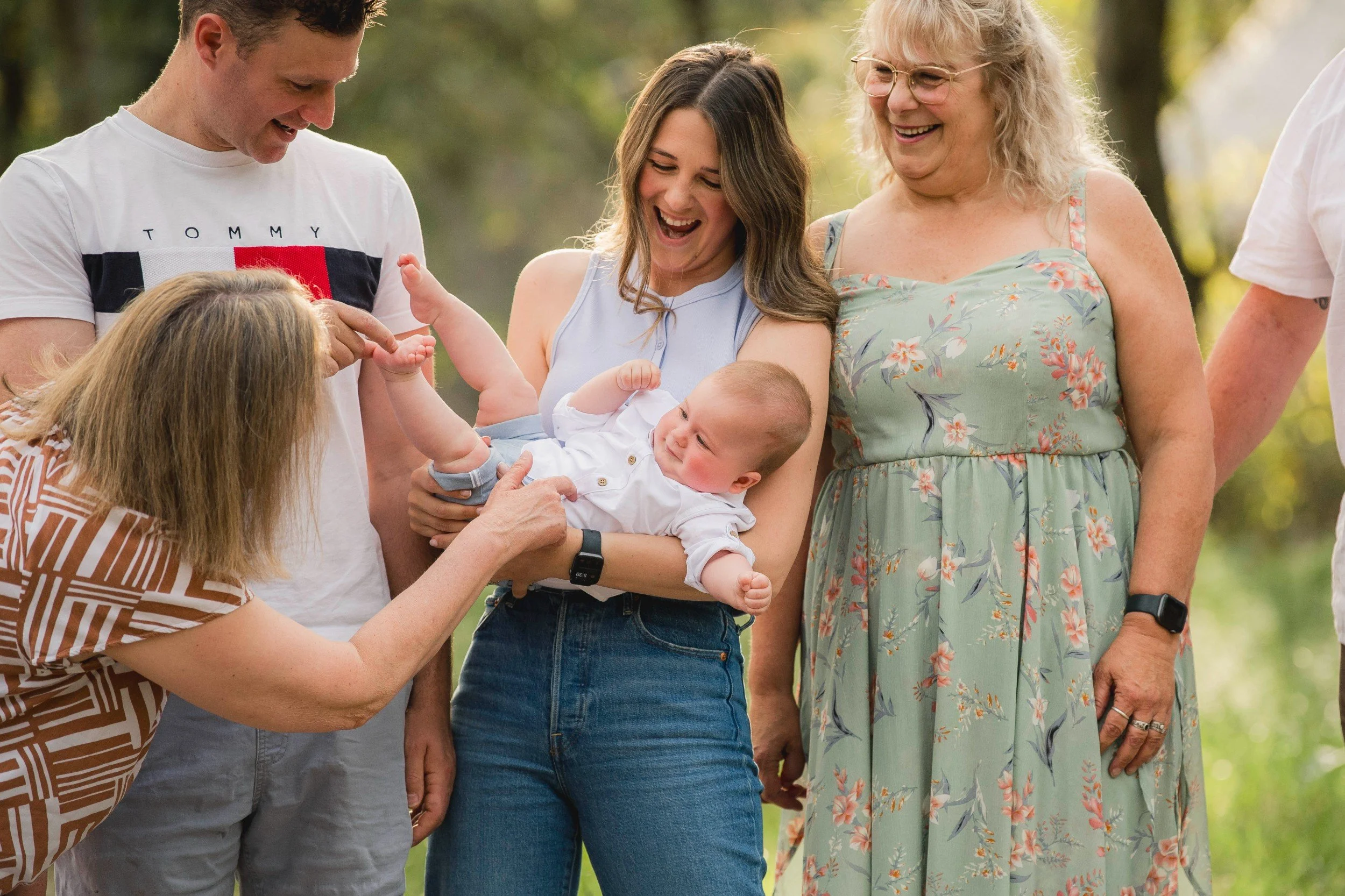 A family gathering outdoors with five adults and two children, one of the children is a baby being held by a woman, everyone is smiling and looking happy in a park-like setting with trees.