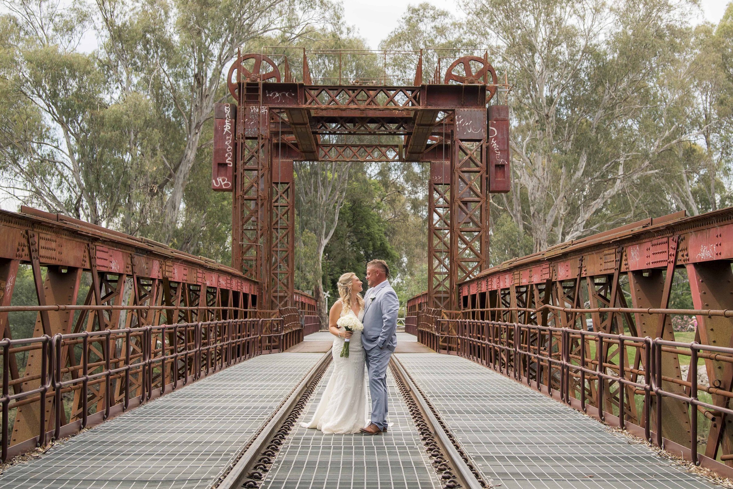 A bride and groom standing close together on a red metal bridge with trees in the background, wedding attire, the bride in a white wedding dress holding a bouquet, and the groom in a light gray suit.