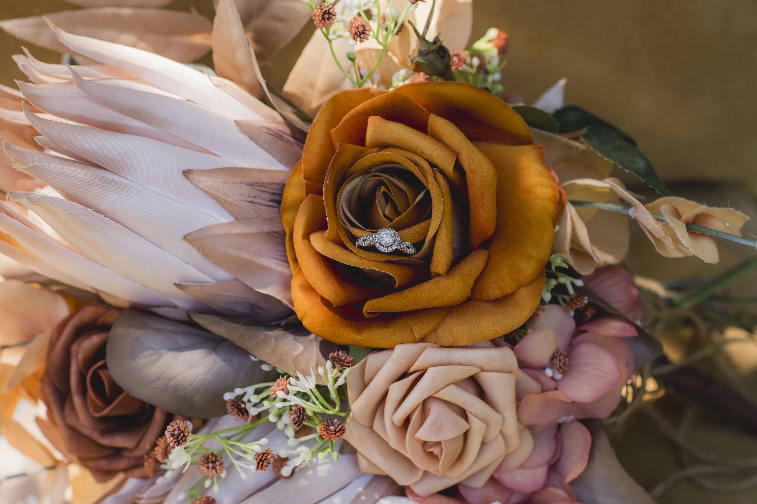 Close-up of a bouquet with a yellow-orange rose and various dried flowers, including a feather, and an engagement ring nestled inside the rose.