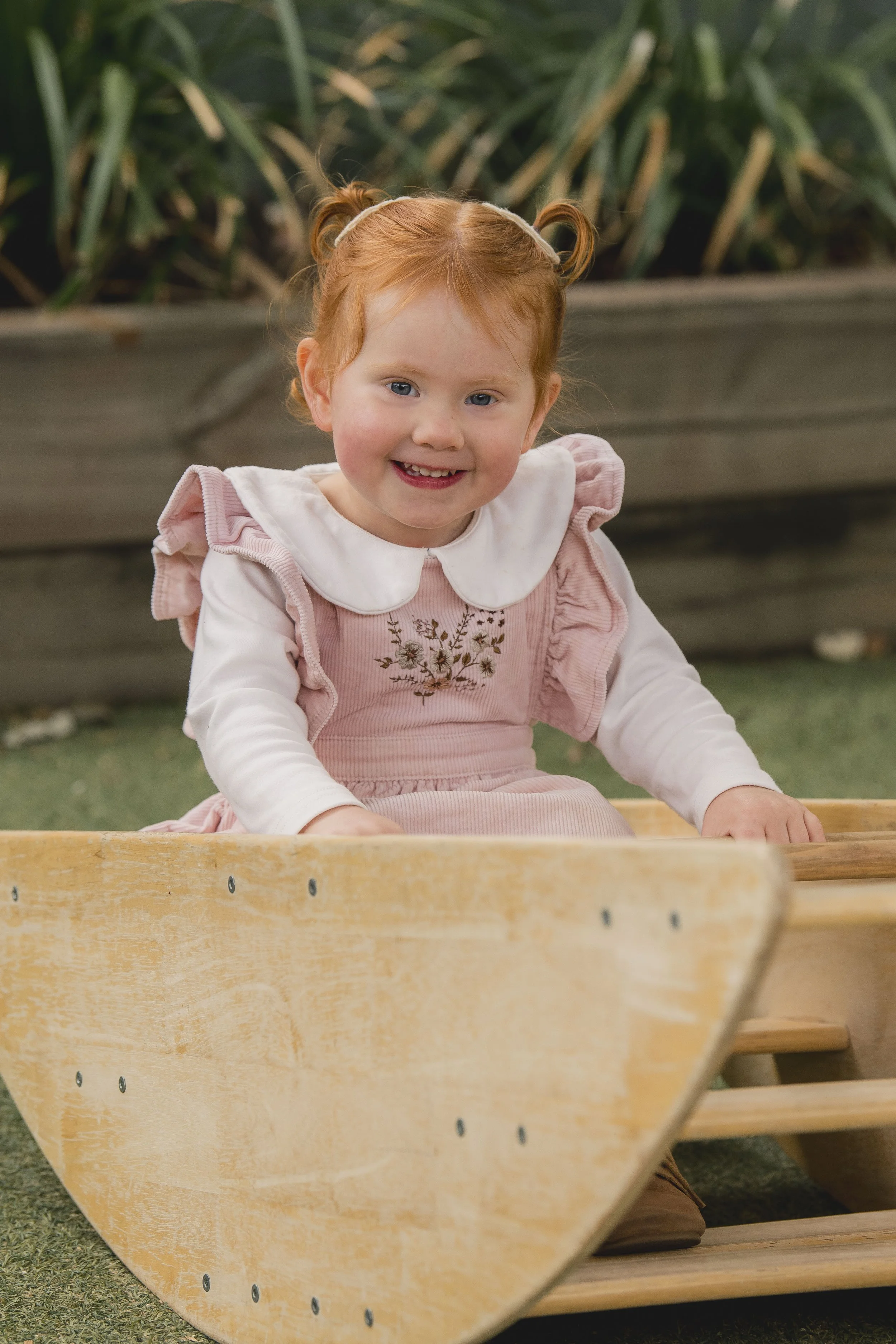 A young girl with red hair styled in pigtails, wearing a pink dress with embroidery, smiling and sitting in a wooden boat outdoors.