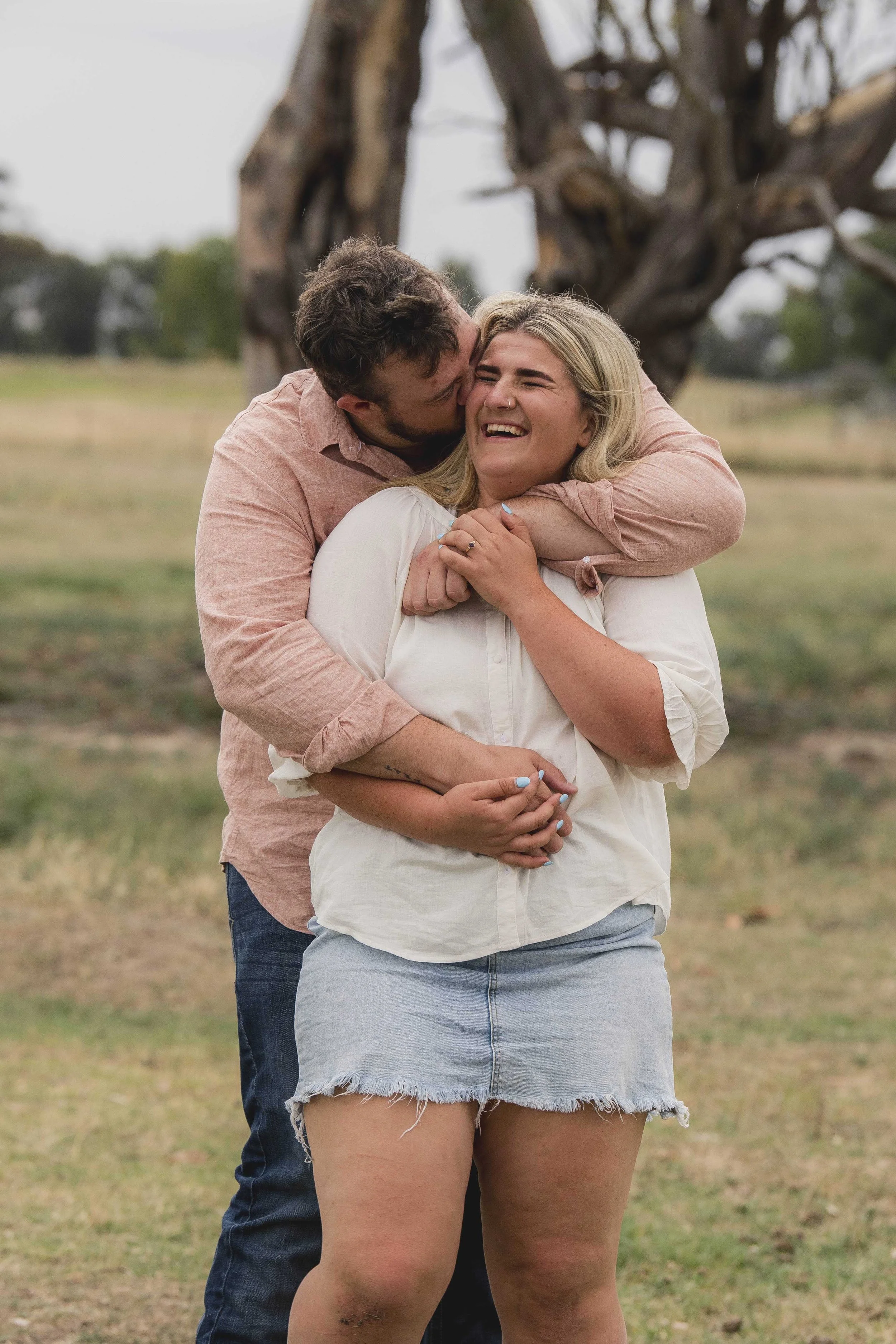 A happy couple embracing outdoors, with a large, twisted tree in the background. The man is kissing the woman's cheek, and they are both smiling. The woman is wearing a white shirt and shorts, and the man is wearing a pink shirt and dark jeans.