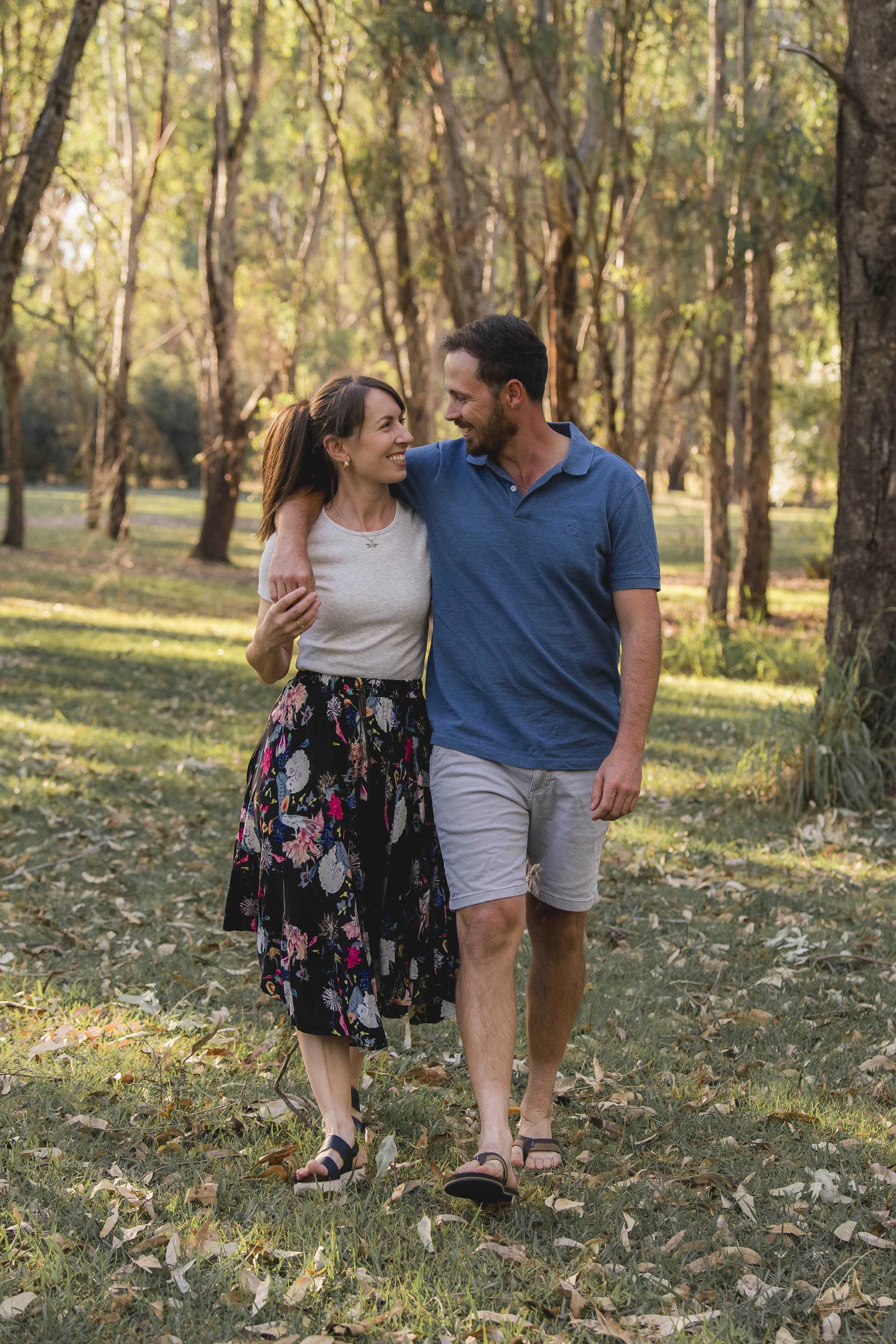 A smiling couple walking together in a park surrounded by trees on a sunny day. The woman has brown hair and is wearing a white shirt and floral skirt, and the man has dark hair and is wearing a blue polo shirt and light shorts.