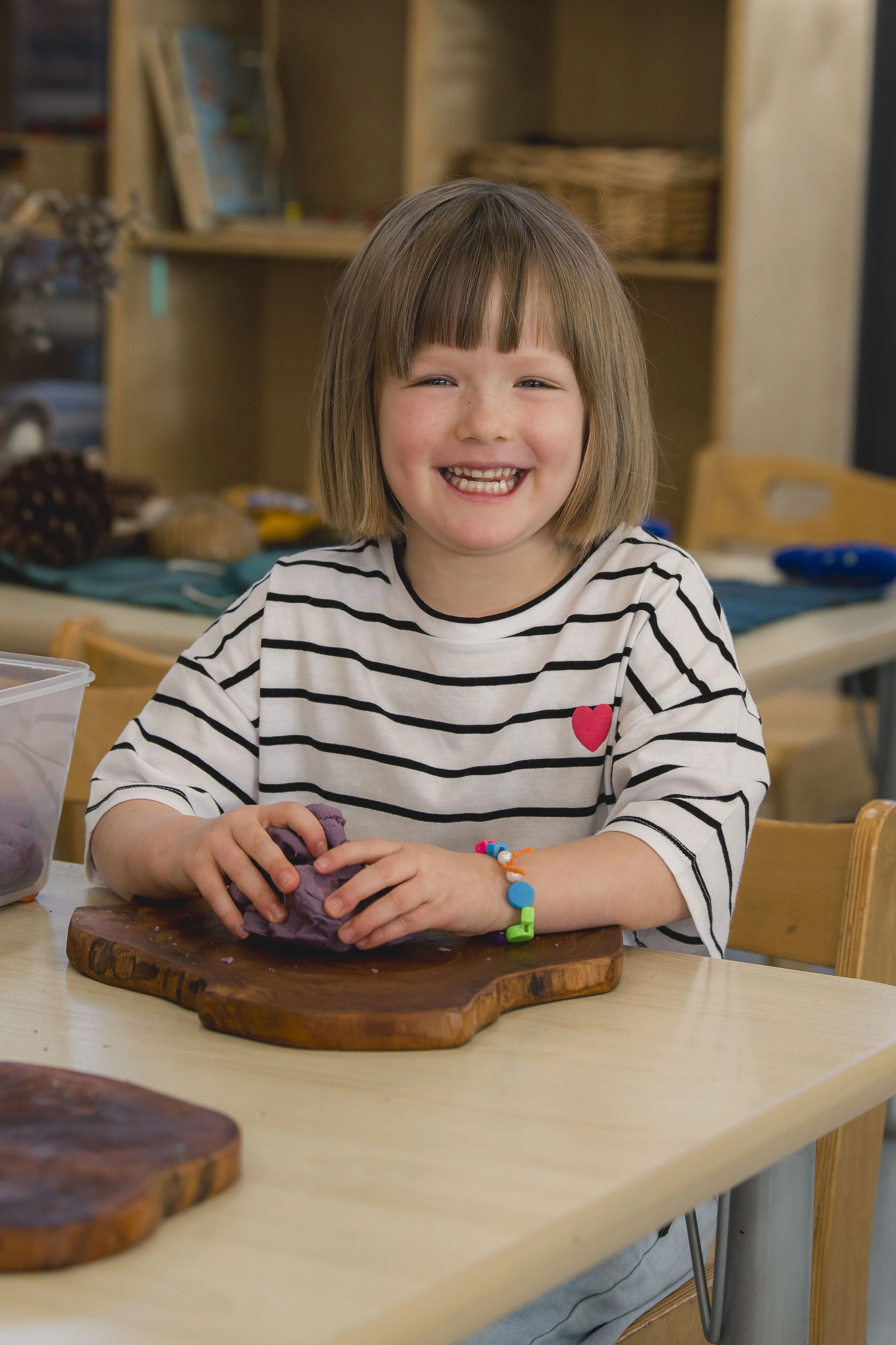 A young girl with shoulder-length blonde hair, wearing a black and white striped shirt with a red heart, smiling while working with purple clay on a wooden cutting board at a table in a classroom or art space.