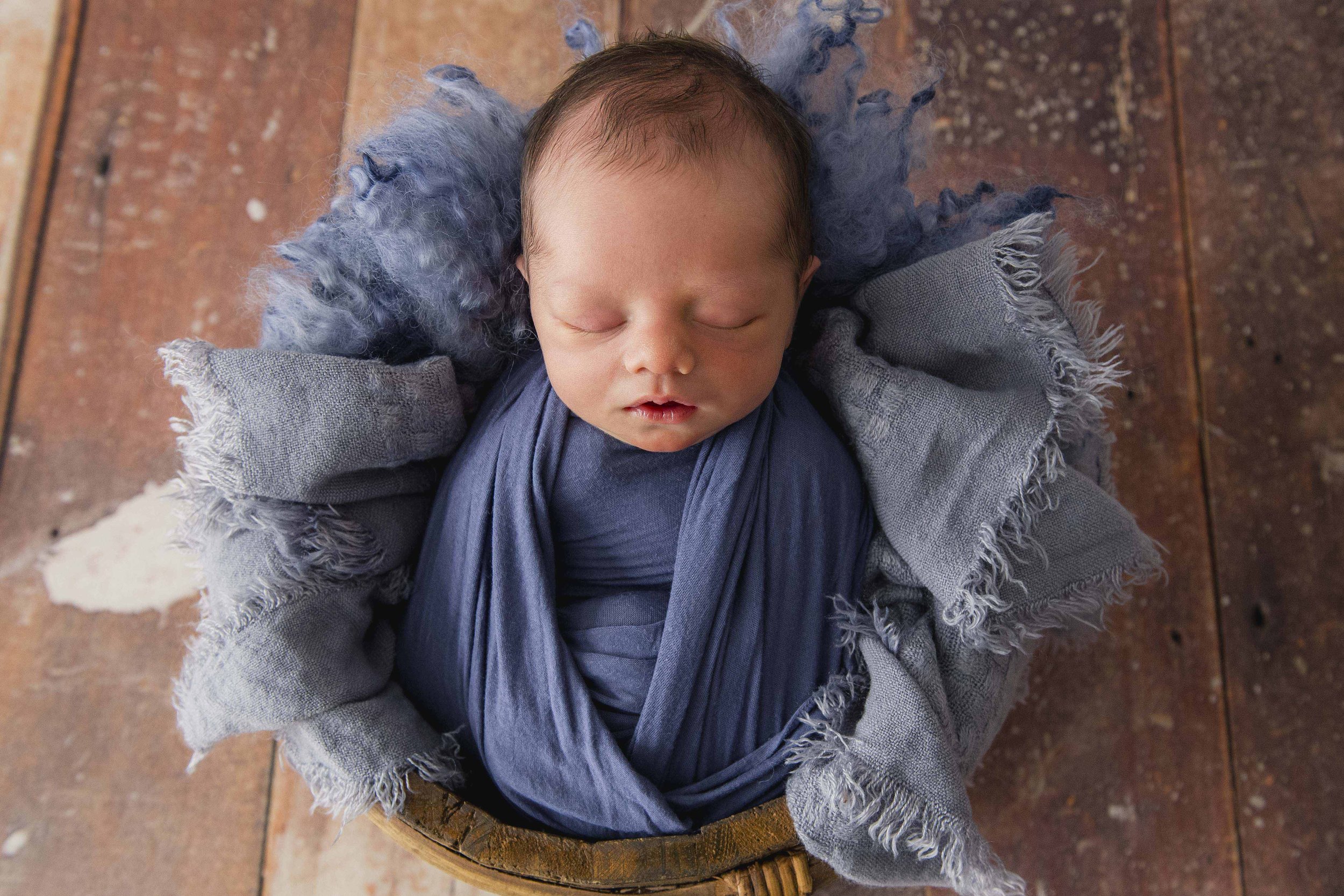 Top-down view of a sleeping baby wrapped in a blue blanket, lying on a wooden surface, with textured gray and fluffy blue and gray fabric around them.