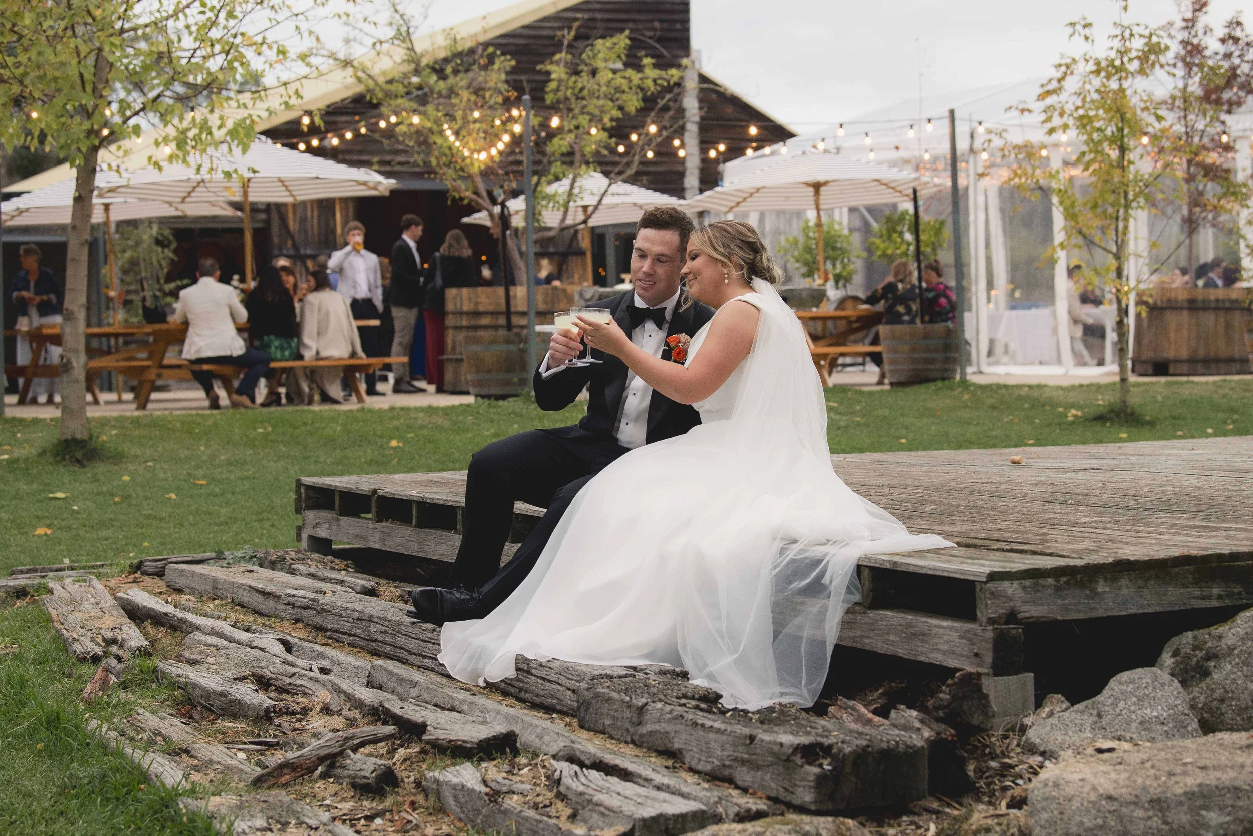 A newlywed couple dressed in wedding attire sitting on a wooden platform outdoors, smiling and holding drinks, at a festive outdoor event with string lights, umbrellas, and guests in the background.