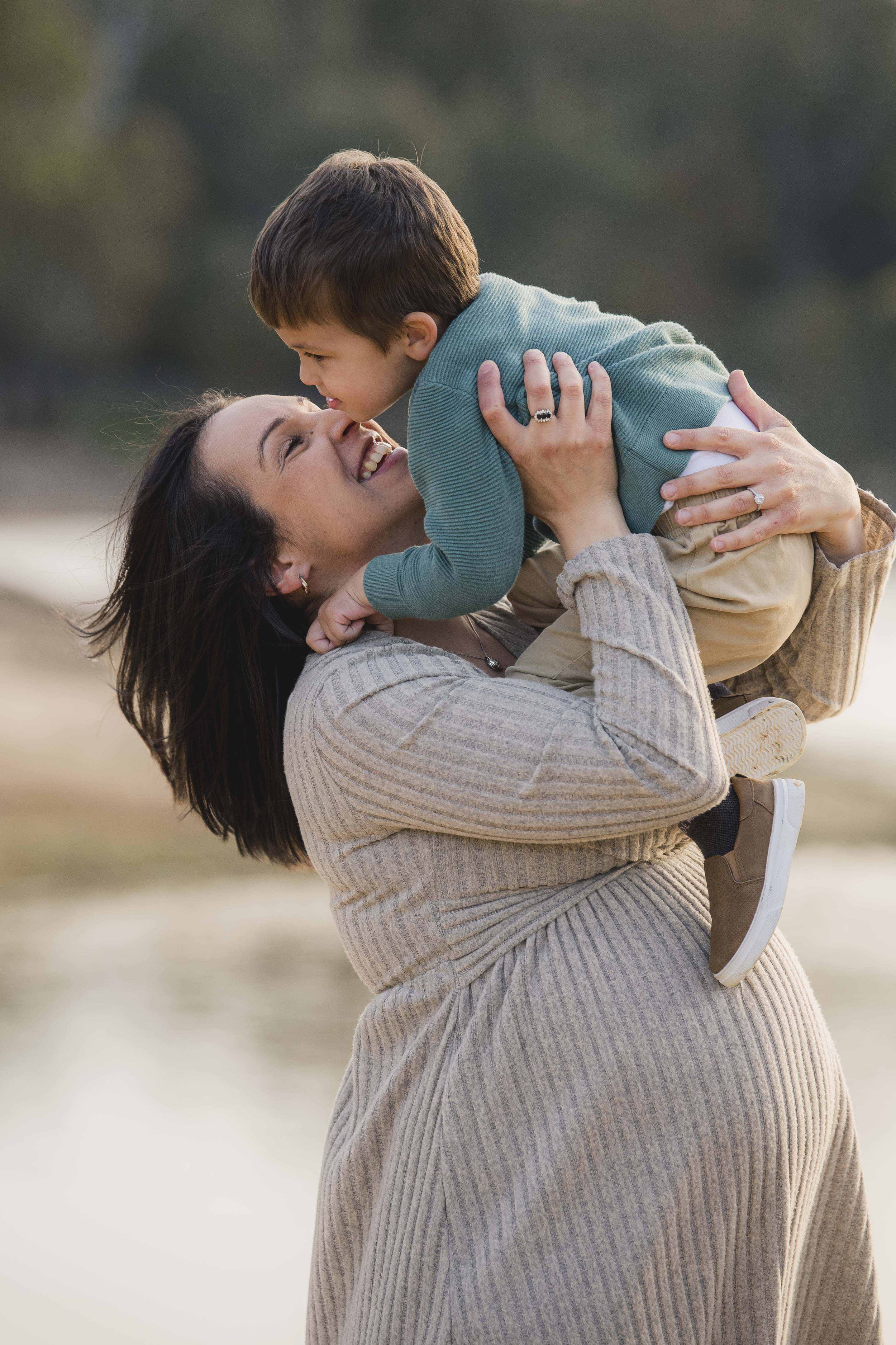 A woman lifting a young boy into the air outdoors, both smiling and laughing, with a blurred natural background.