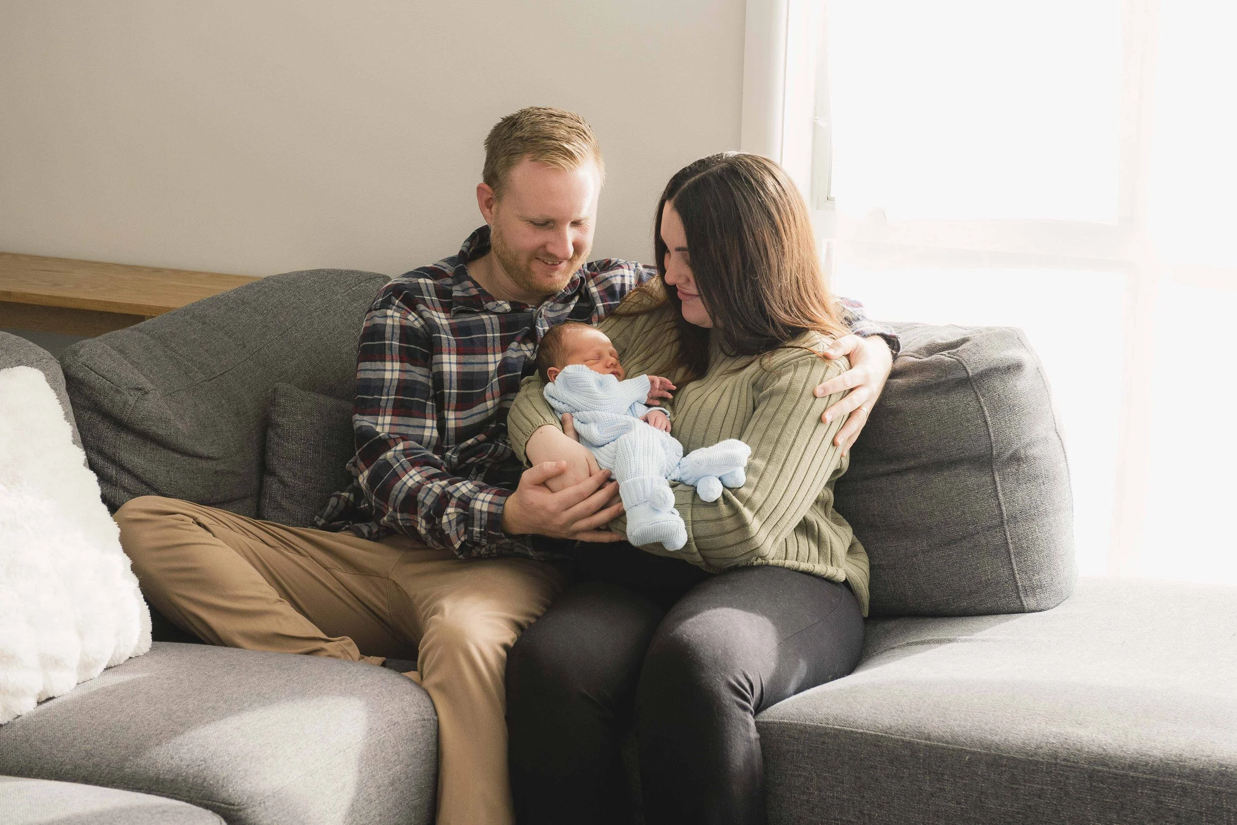 A happy family sitting on a gray sofa in a bright living room, holding and looking at a newborn baby wrapped in a blue blanket.