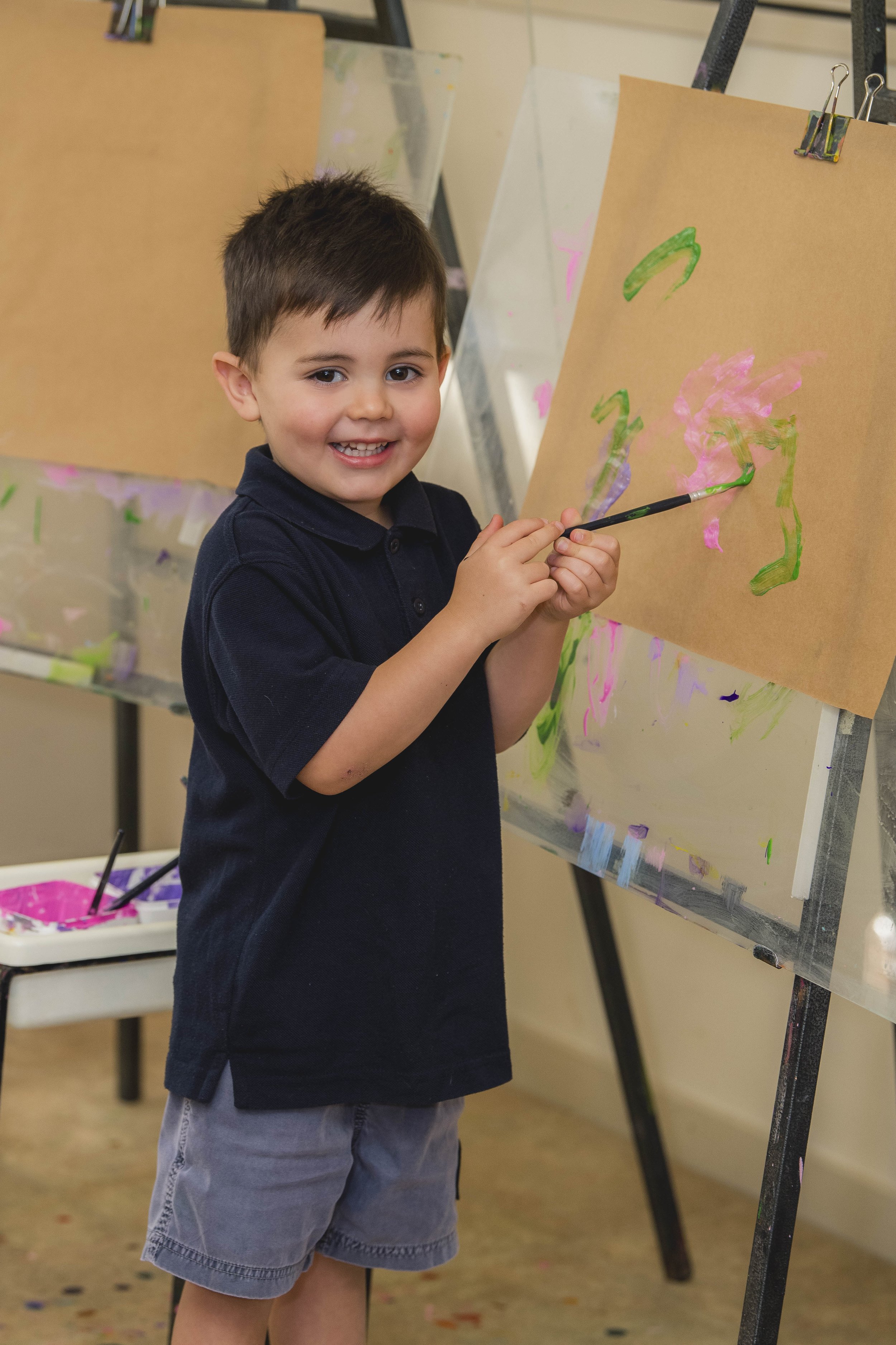 A young boy with short brown hair and a big smile, wearing a navy blue polo shirt and gray shorts, is standing in front of an easel with brown paper, painting with a brush on a canvas. The background shows a table with pink paint containers and other art supplies, indicating an art classroom or studio.