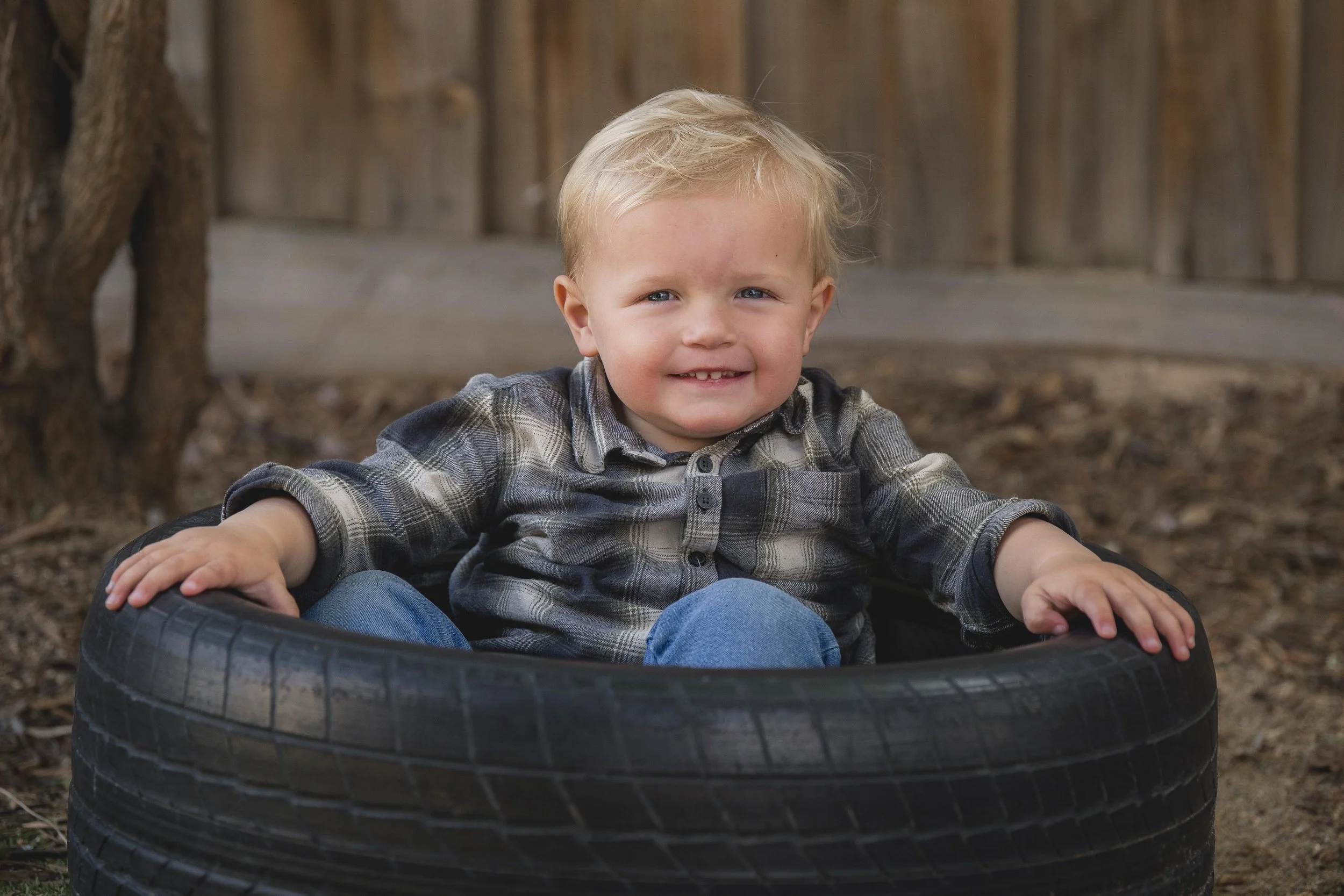 A young boy with blonde hair and blue eyes, smiling, sitting inside a black tire in an outdoor area with a wooden fence in the background.