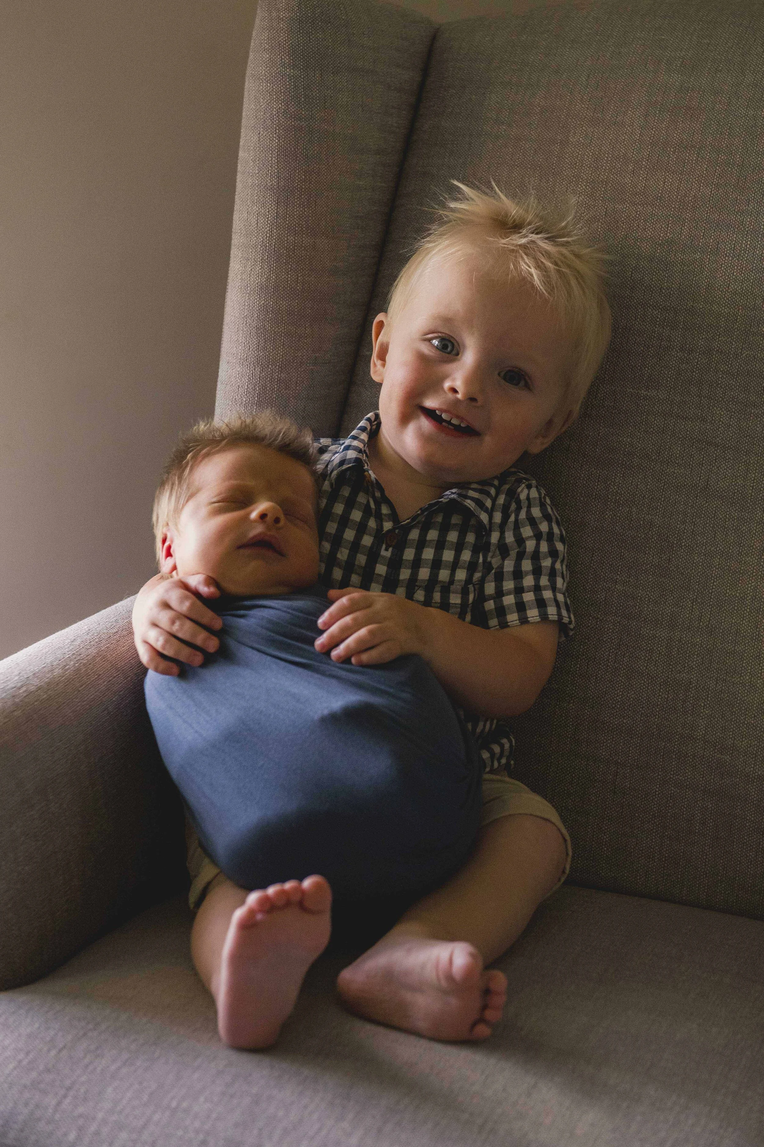 A young boy with blonde hair and a big smile holding a sleeping infant on a gray couch.