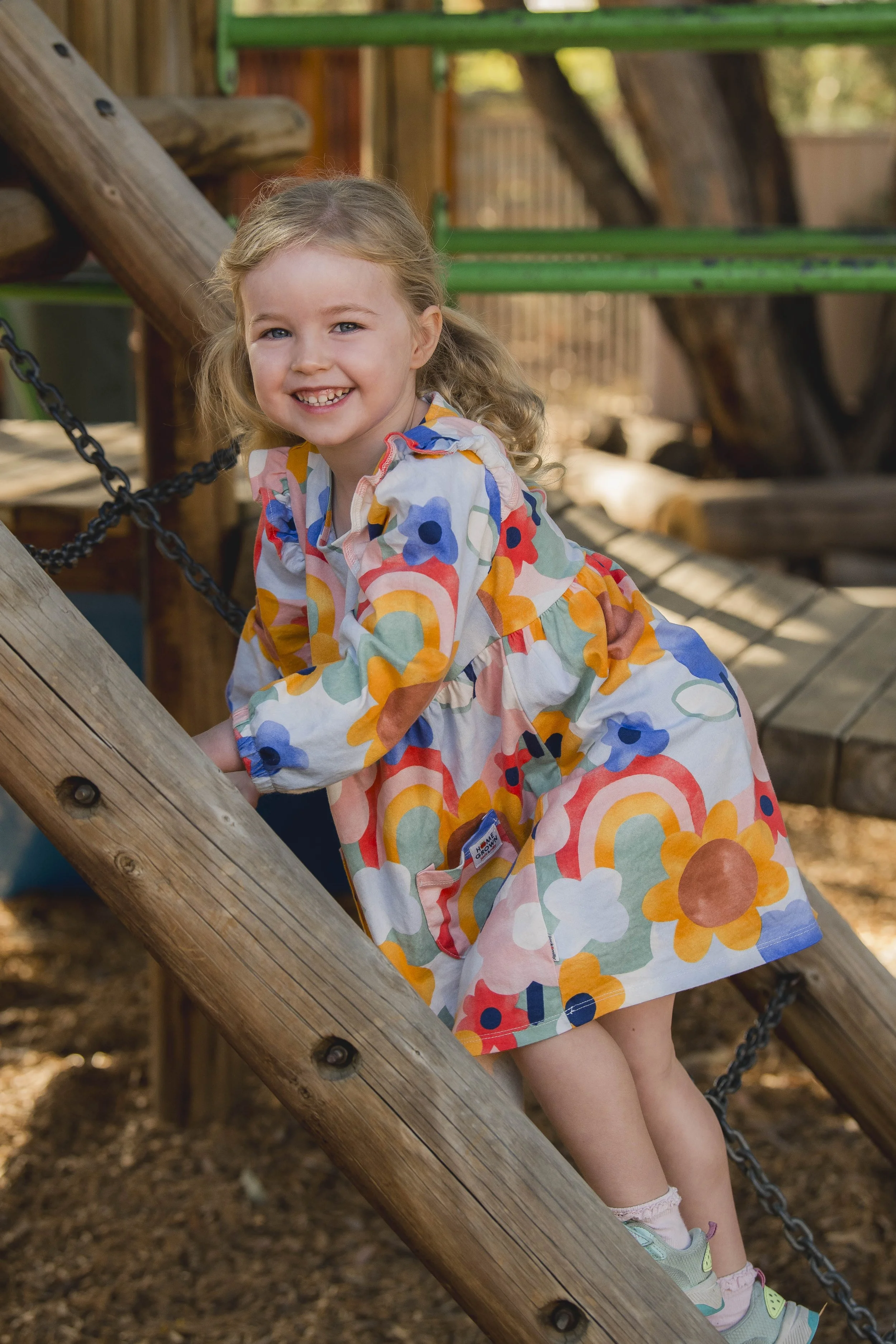 A young girl with curly blonde hair, smiling, wearing a colorful floral dress, playing on a wooden playground structure with green accents, outdoors with trees and dirt ground.