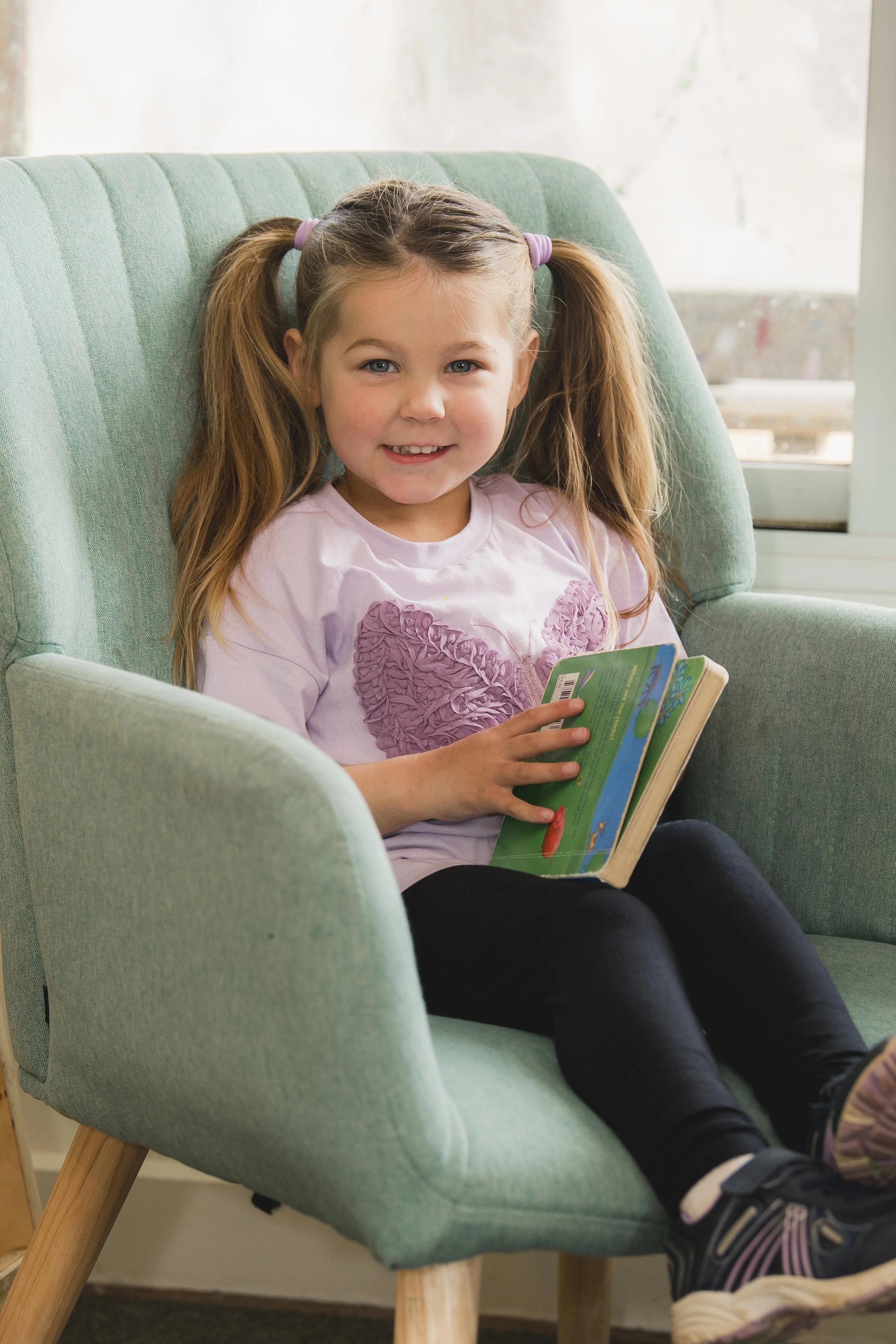 A young girl with pigtails and a pink shirt sitting in a green armchair, smiling and holding a colorful book, near a window.