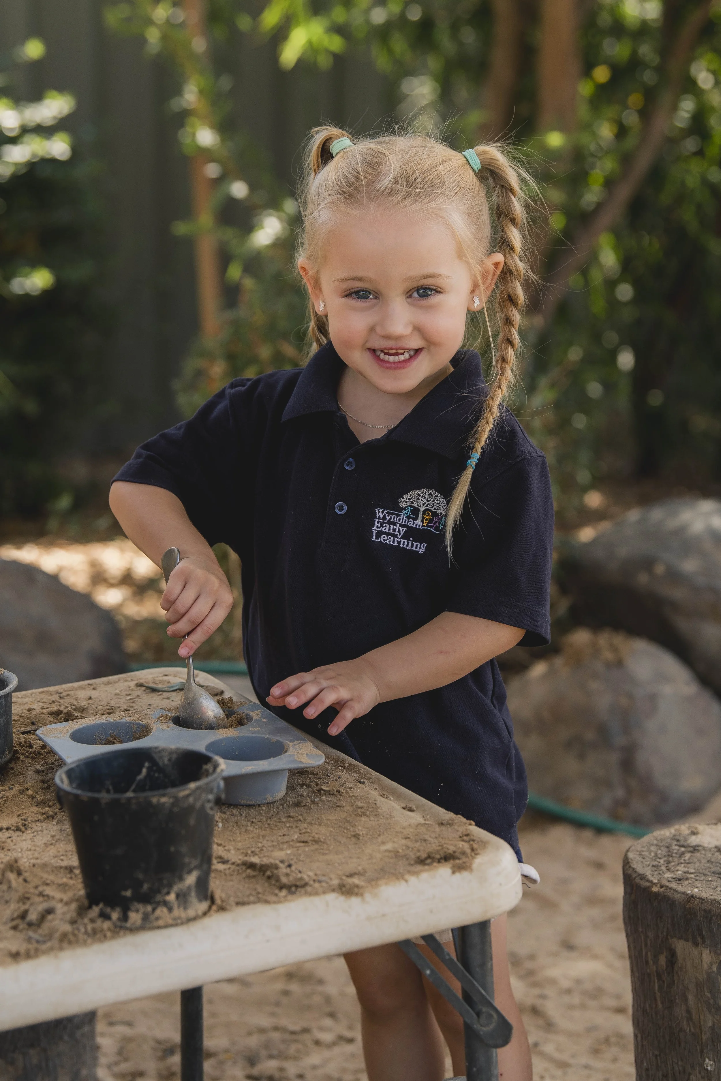 A young girl with blonde hair in pigtails and wearing a dark uniform is playing outside at a sandbox, using a spoon. There are sand molds and cups on the sandbox in front of her, with greenery in the background.