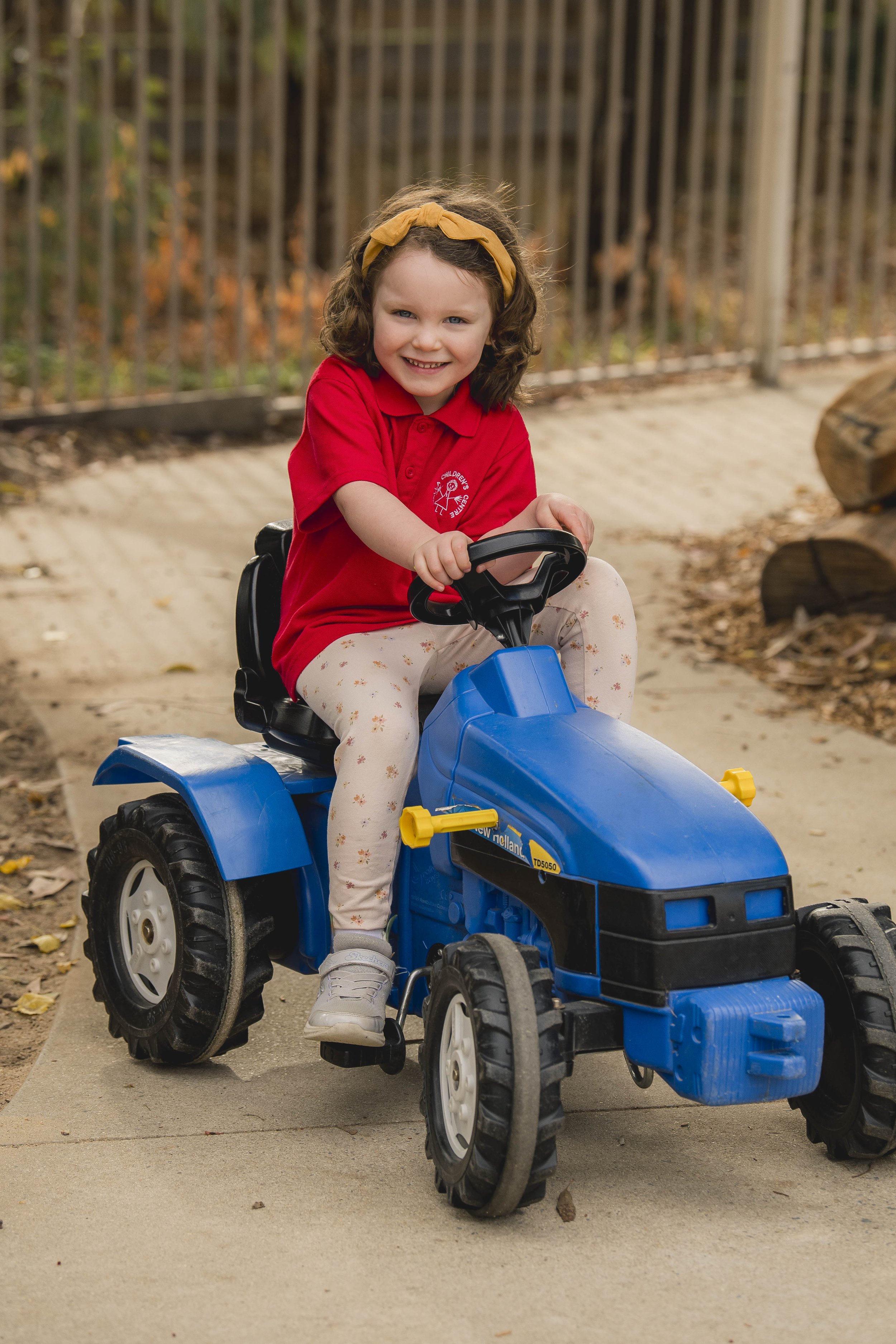 A young girl with curly brown hair, wearing a yellow headband, red shirt, and floral leggings, is riding a blue toy tractor outside on a paved path with fallen leaves and a metal fence in the background.