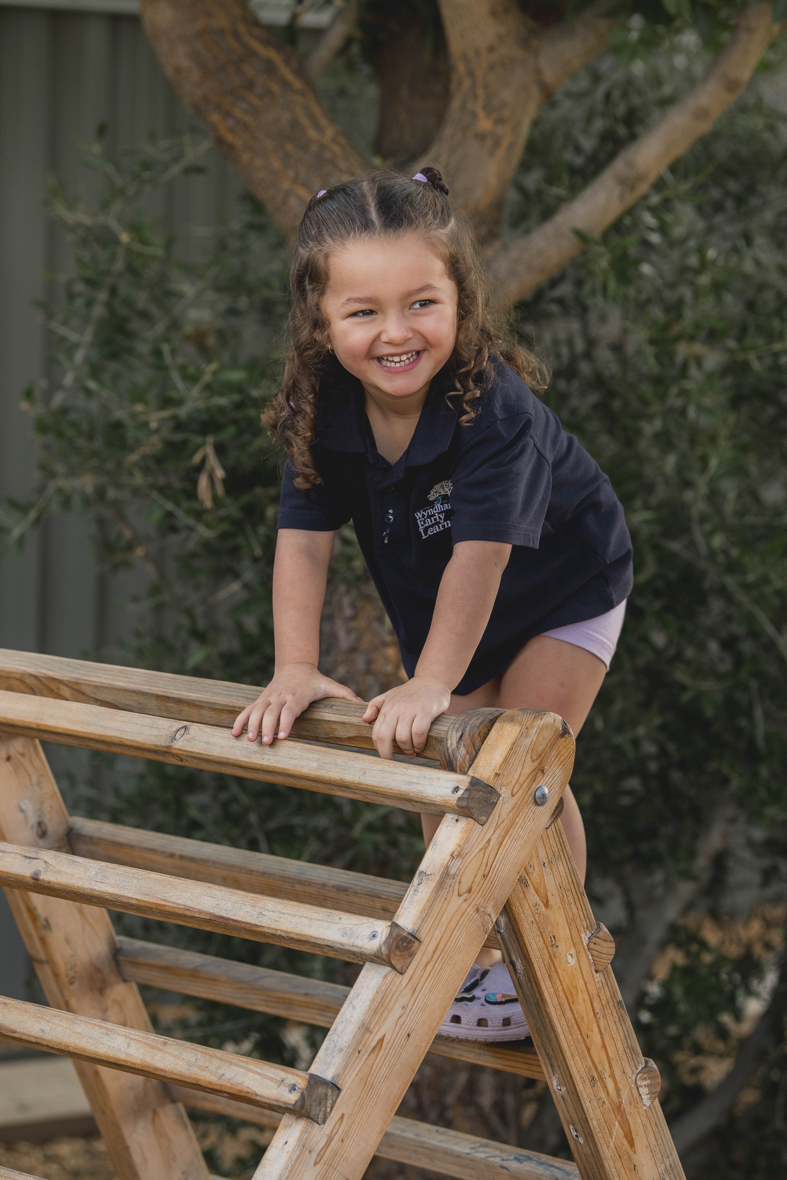 A young girl with curly brown hair in pigtails, wearing a navy blue polo shirt with a logo, is climbing on a wooden structure outdoors, smiling and looking to her left, with a large tree in the background.