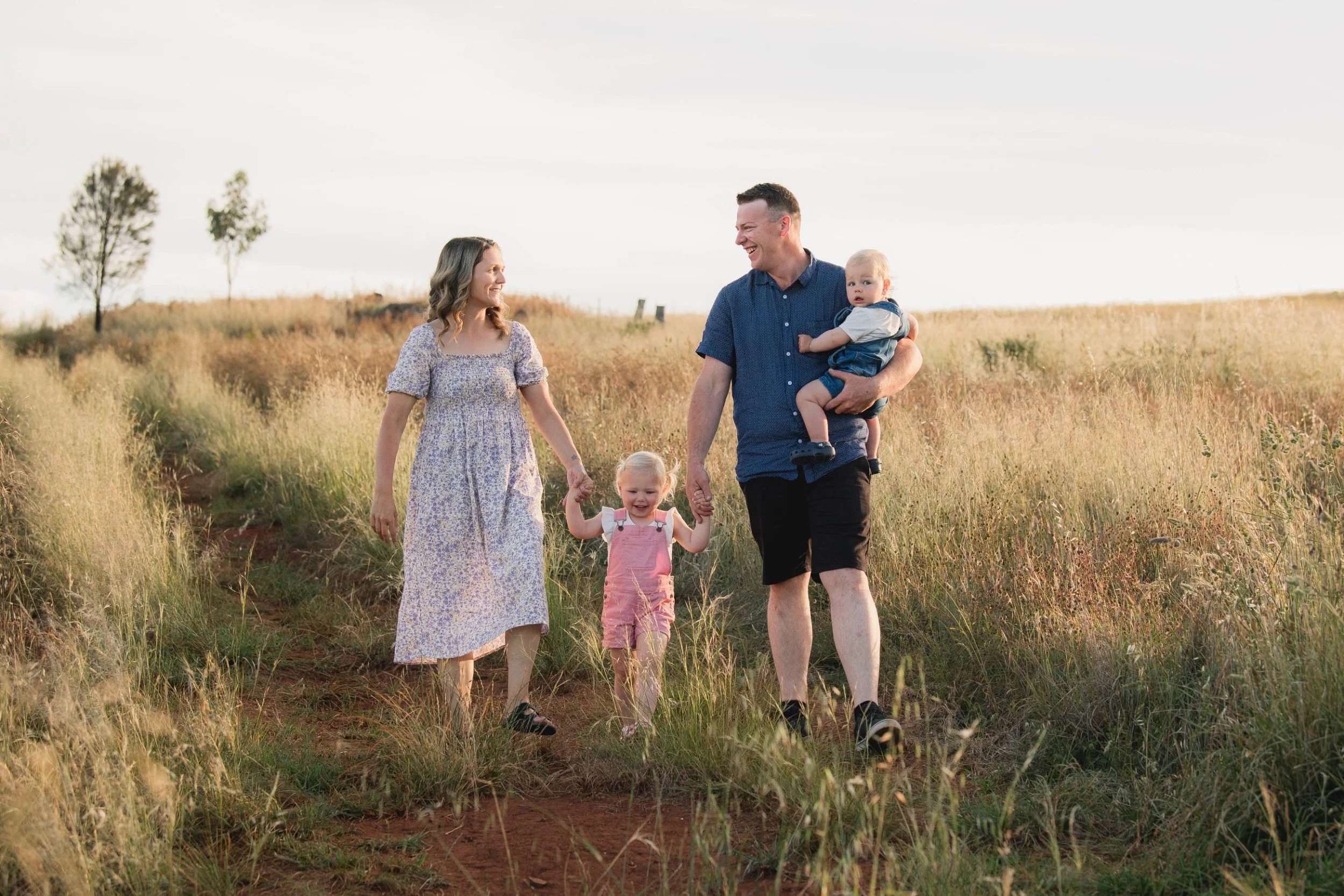 A family of four walking through a grassy field in the late afternoon, smiling and holding hands, with two trees in the background.