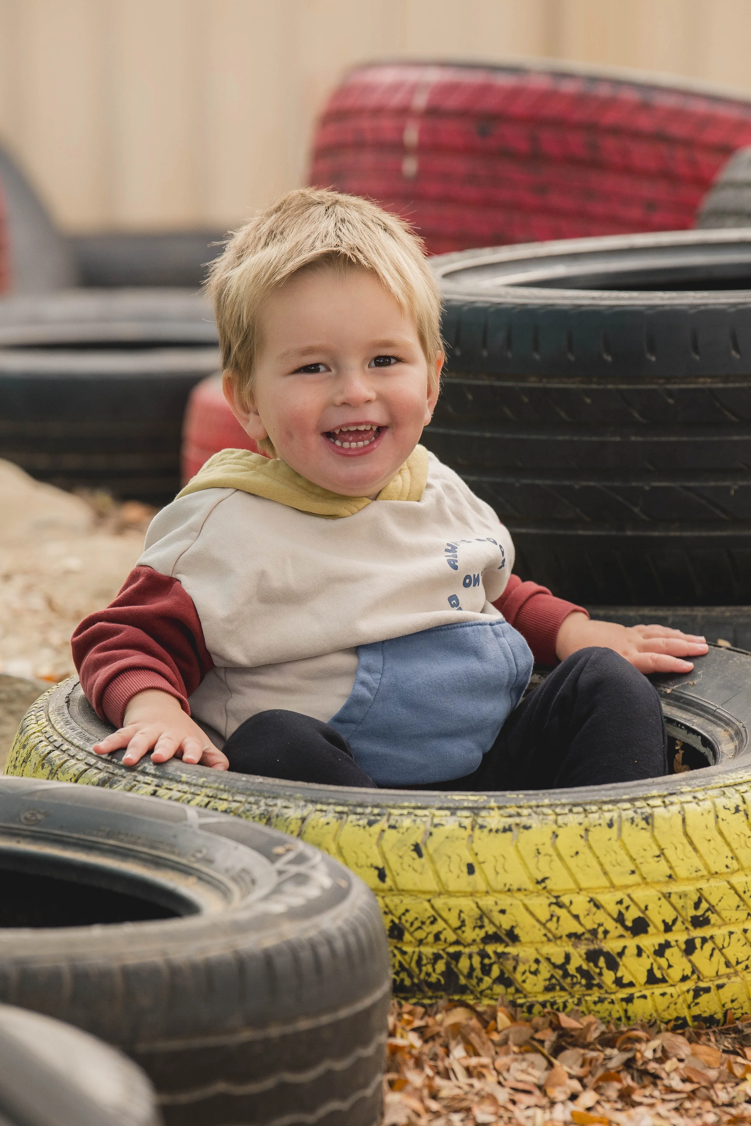 A smiling young boy with blonde hair sitting inside a stack of old tires outdoors, surrounded by fallen autumn leaves.