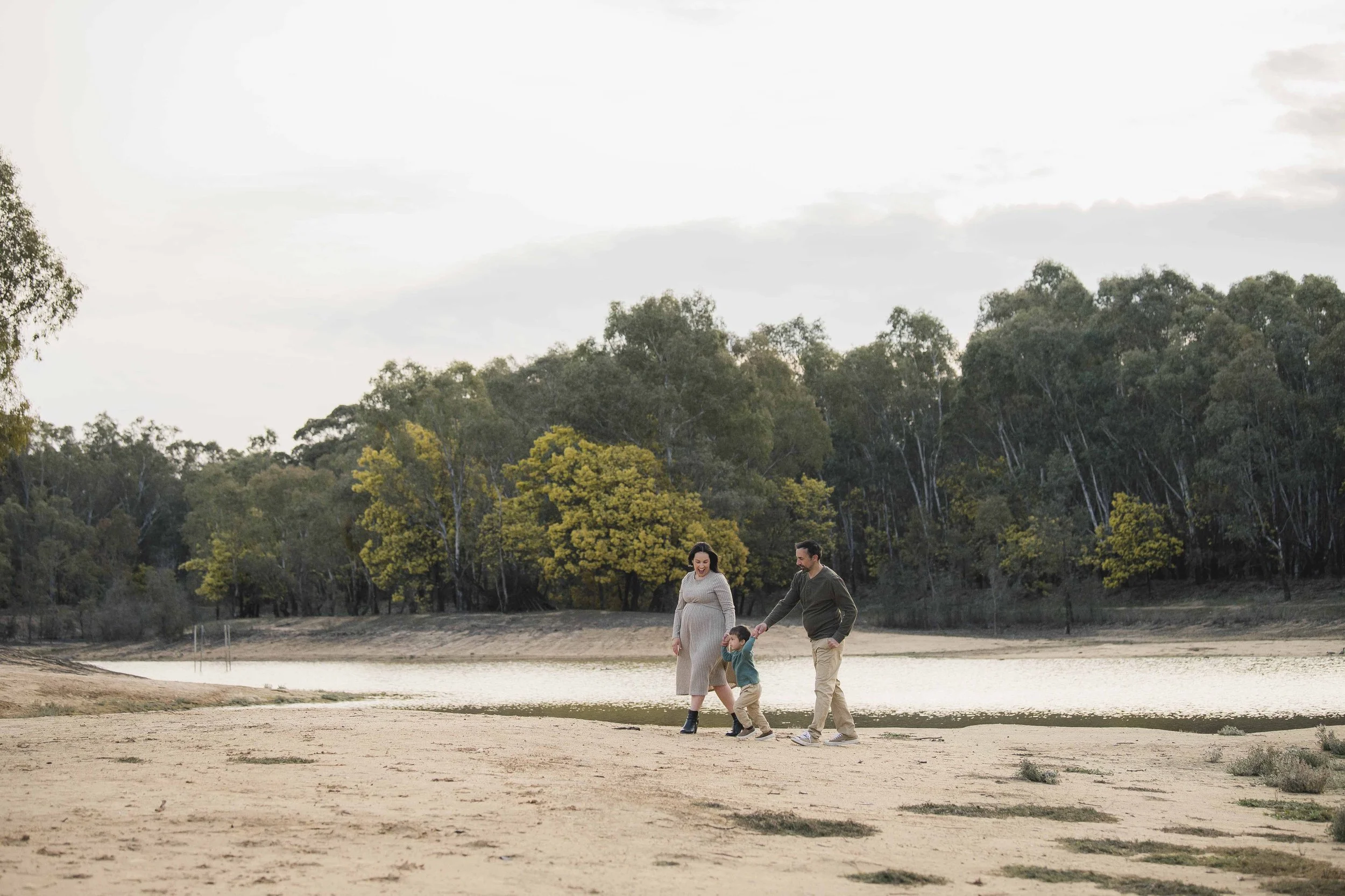 A family of three walking by a body of water with tall trees in the background, during late afternoon or early evening.