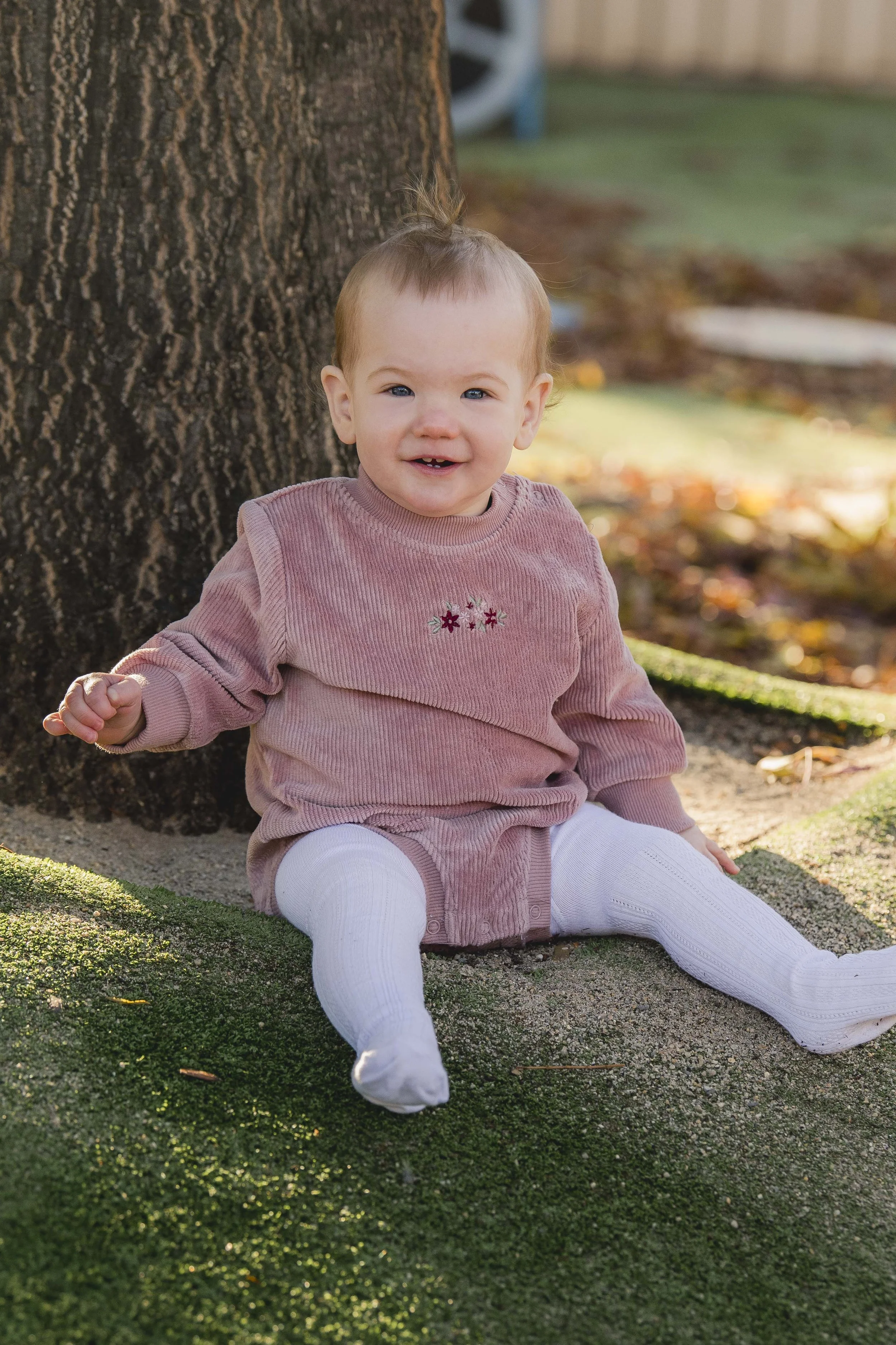 A smiling toddler girl with blue eyes and light brown hair in a small ponytail, sitting outdoors on a patch of artificial grass, leaning against a tree, wearing a pink long-sleeve top with embroidery, white tights, and white shoes, with a background 