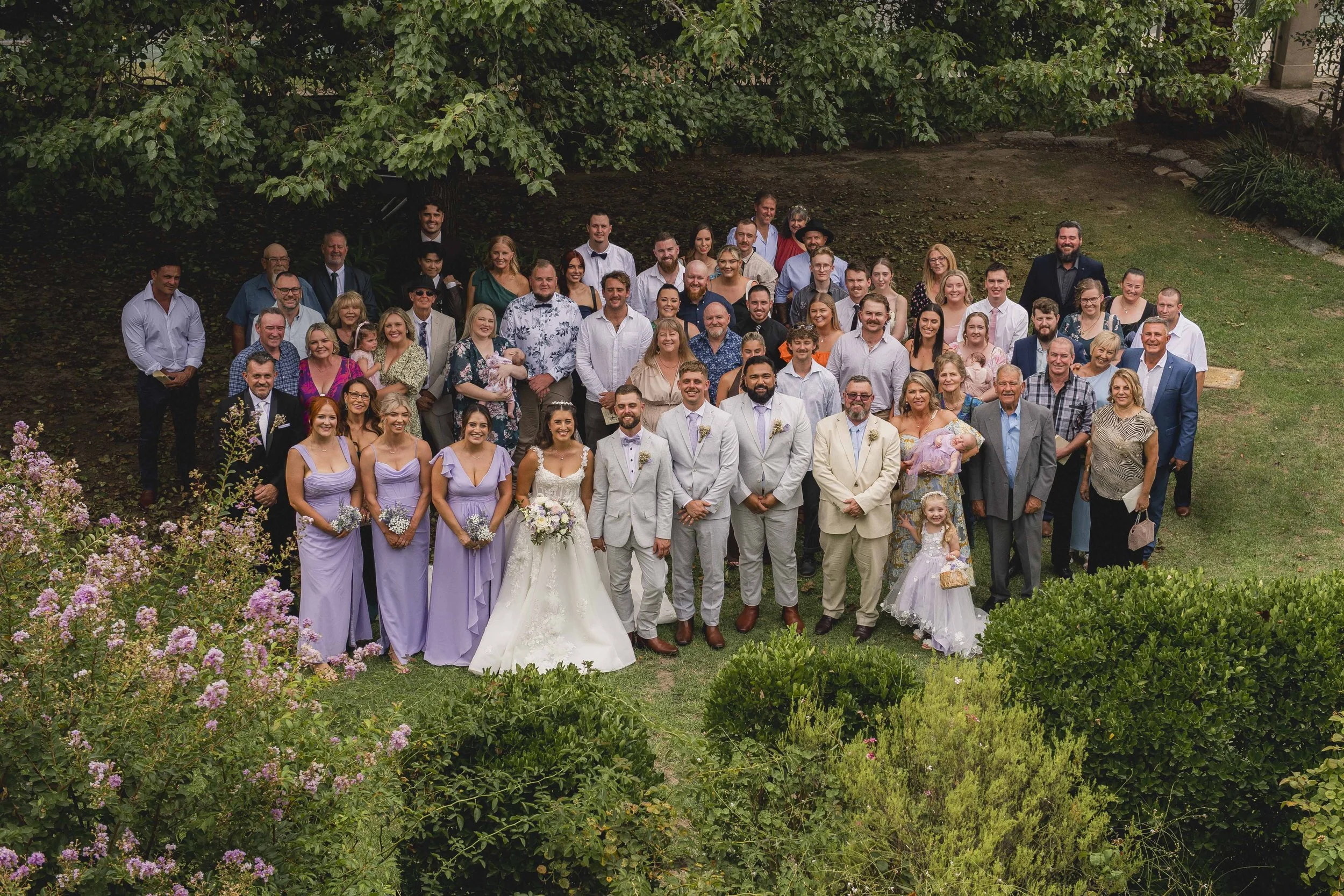 A large group of people gathered outdoors for a wedding photo, standing on grass under a large tree with pink flowers in the foreground.