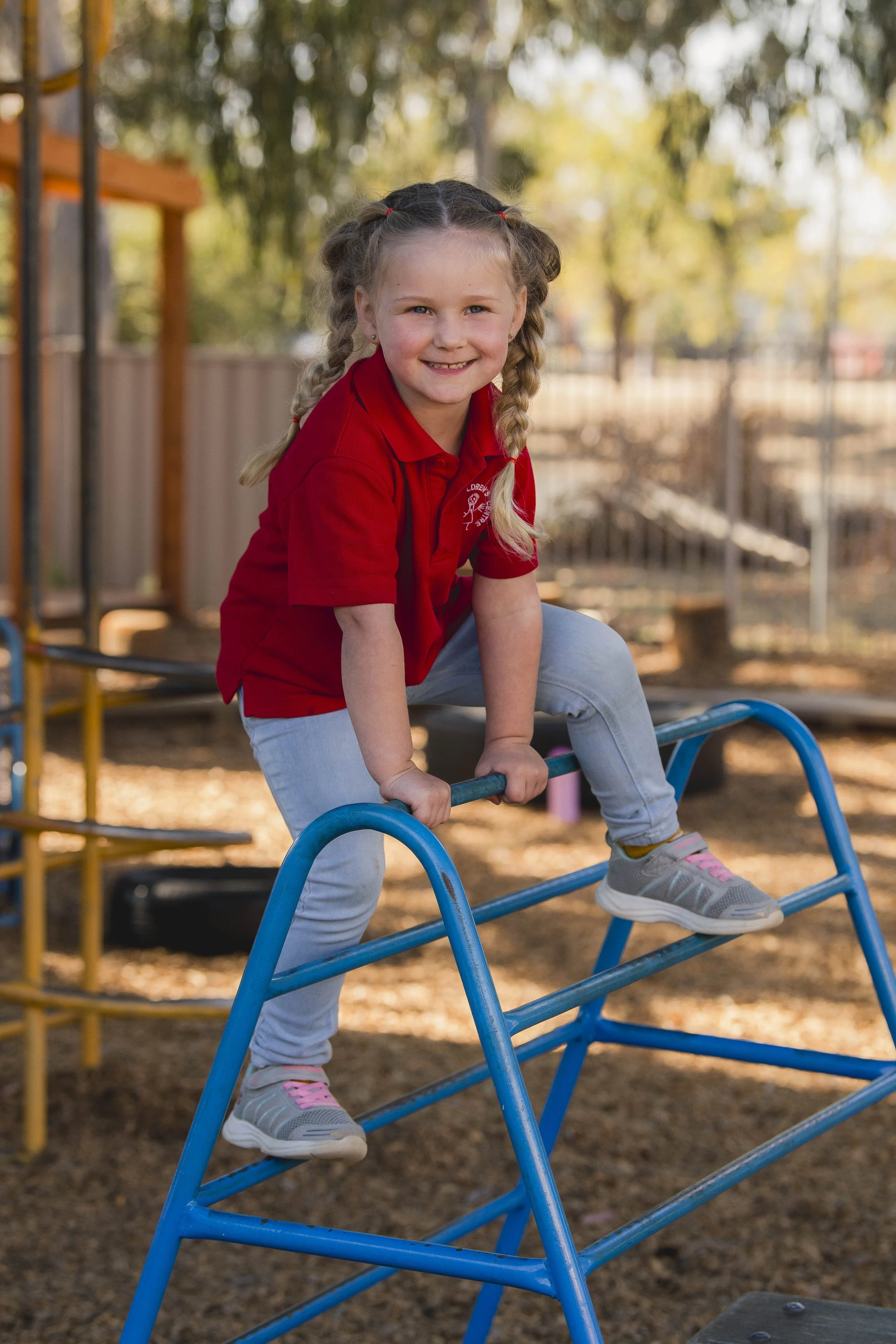 A young girl with braided hair, wearing a red polo shirt and gray pants, is playing on a playground structure in an outdoor park. She is smiling and holding onto a blue metal curved ladder, with trees and a wooden fence in the background.