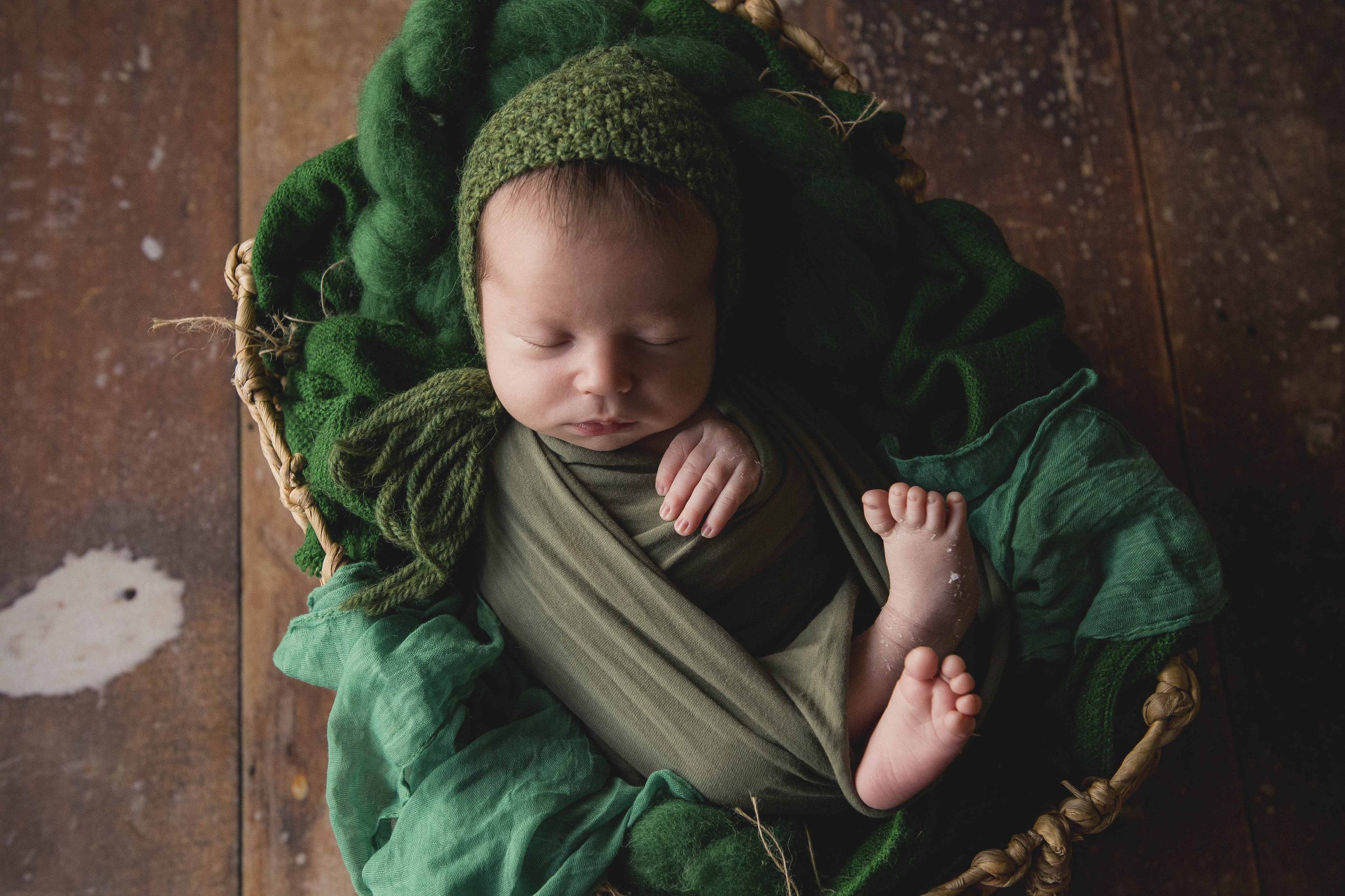 A sleeping baby dressed in green lying on a wicker basket on a tiled floor.