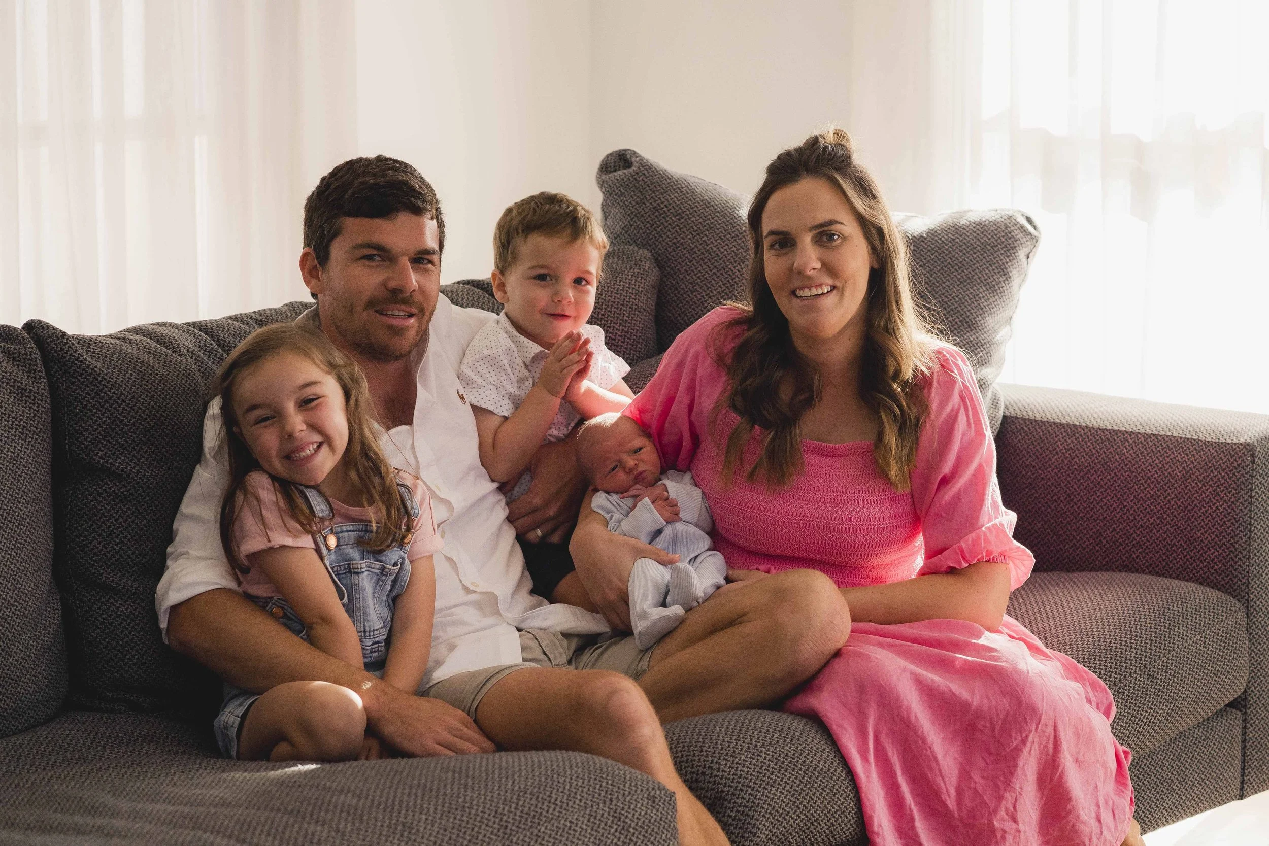 A family of six sitting on a dark gray couch in a bright living room, including a man, a woman, two young girls, a young boy, and a newborn baby.