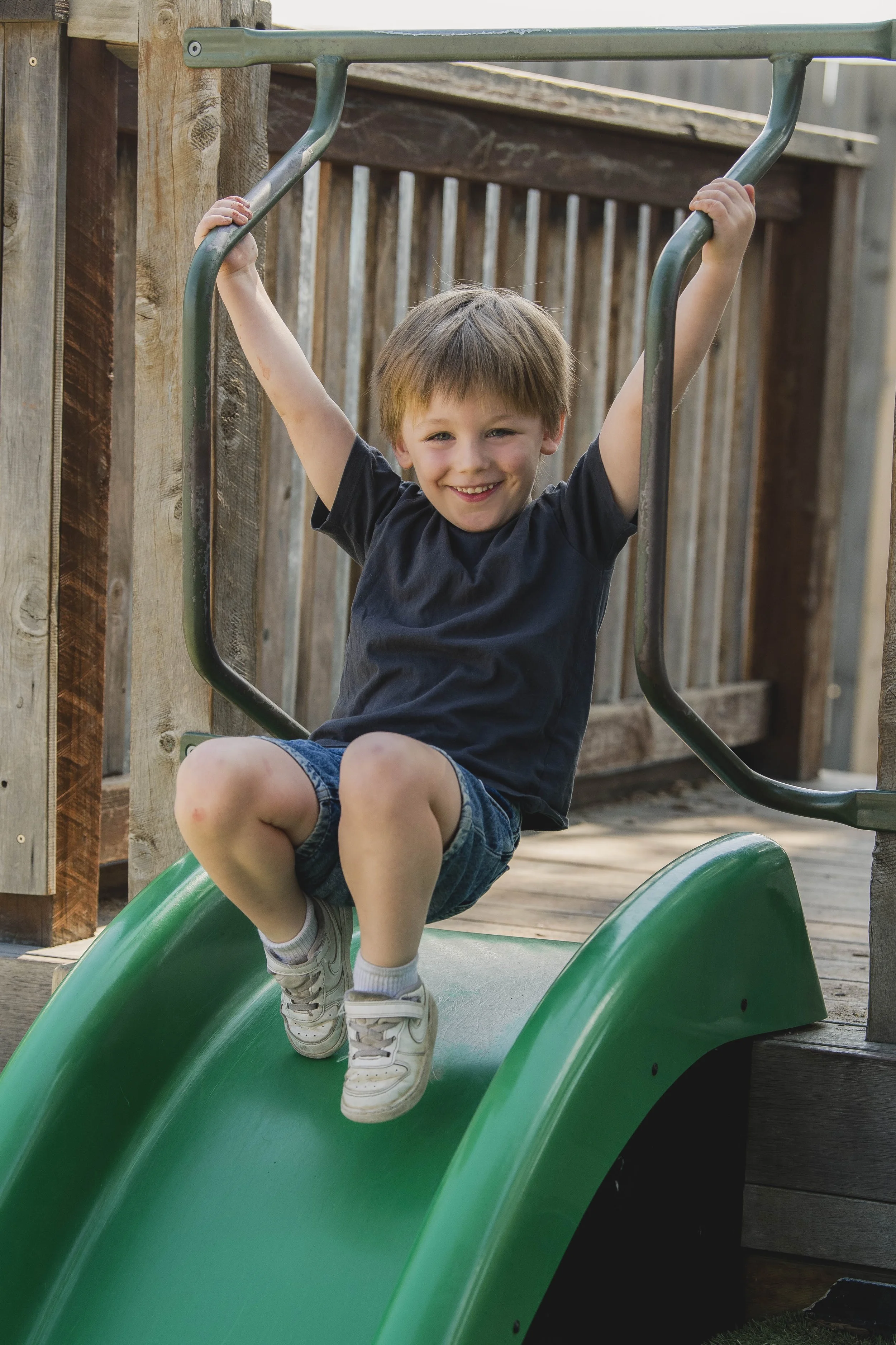 A young boy with brown hair, wearing a black T-shirt, denim shorts, white socks, and sneakers, is sitting on a green slide outdoors. He is smiling and holding onto the metal bars at the top of the slide, with a wooden fence in the background.