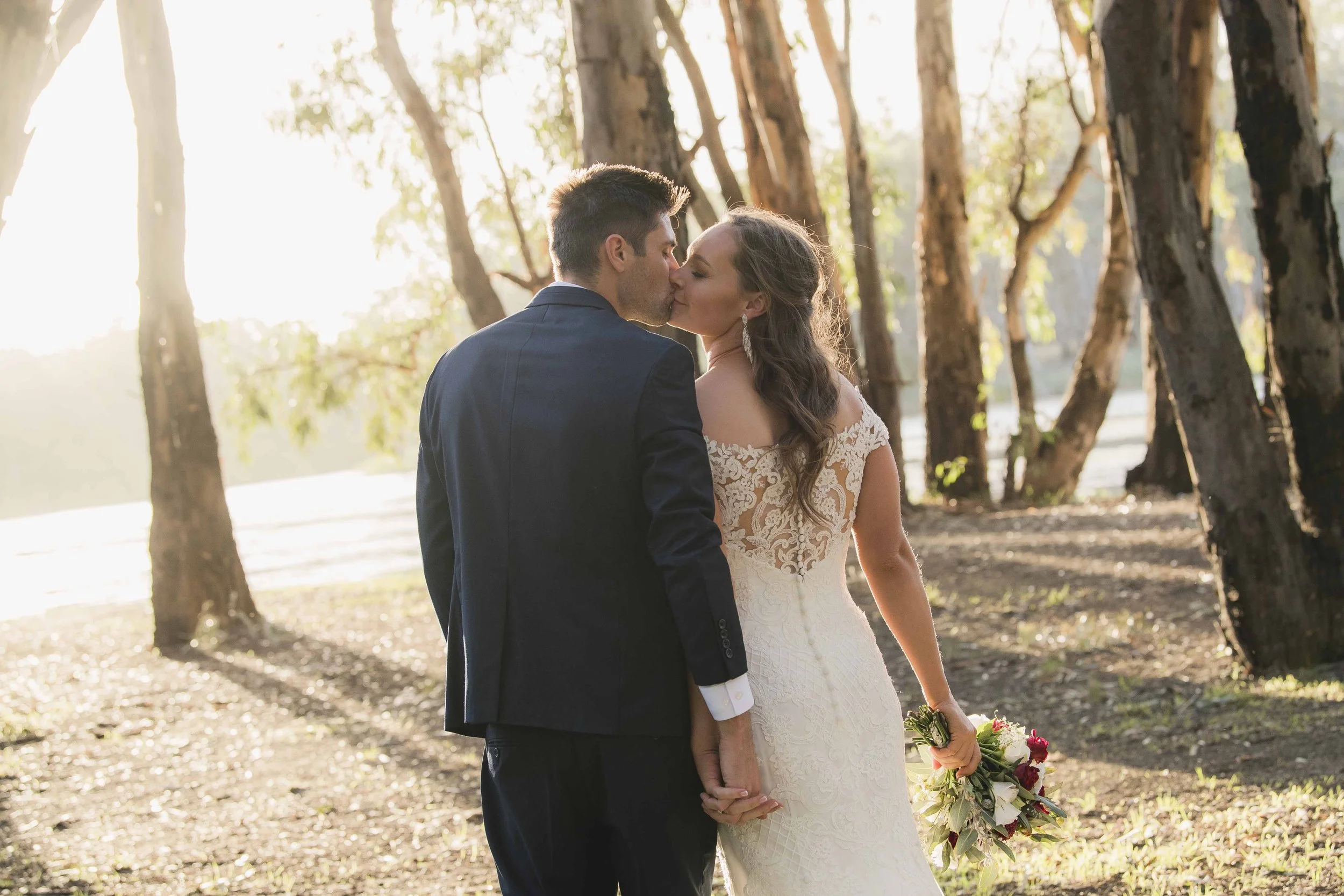 A bride and groom kiss outdoors among trees during a wedding ceremony, with sunlight filtering through the branches.