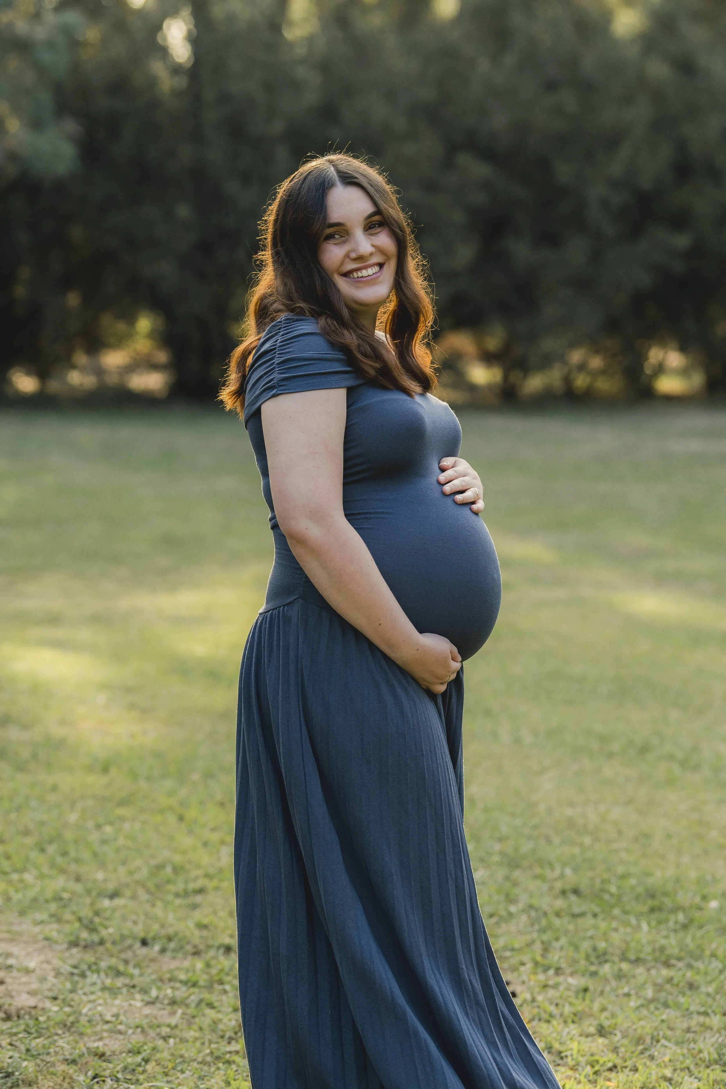A pregnant woman in a blue dress standing outdoors on a grassy field, smiling and holding her belly with one hand, with trees in the background during sunset.