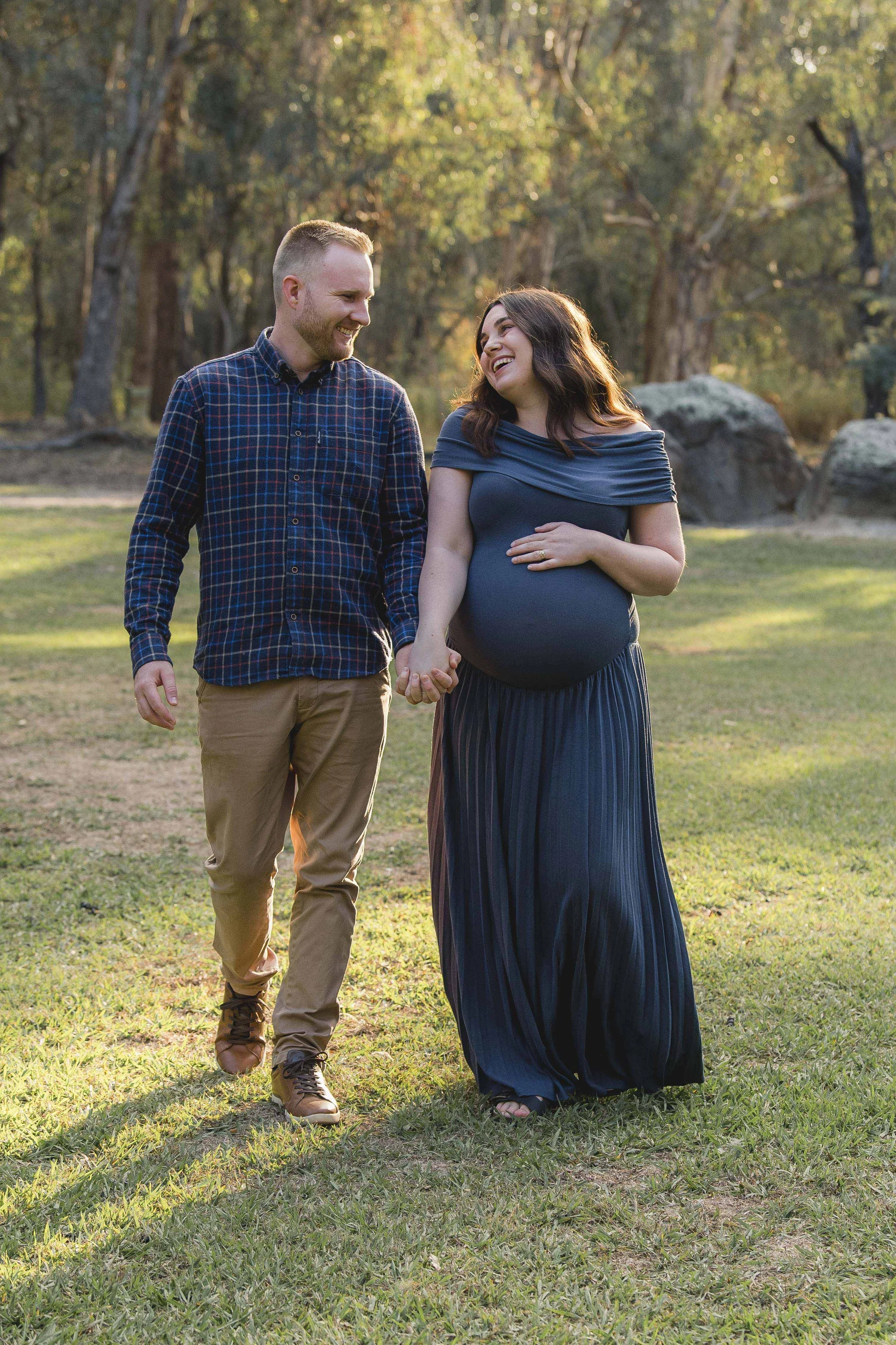 A happy couple walking hand in hand outdoors in a park during sunset. The woman is pregnant, wearing a long dark dress, and the man is casually dressed in a plaid shirt and khaki pants. They are smiling and looking at each other, with trees and large