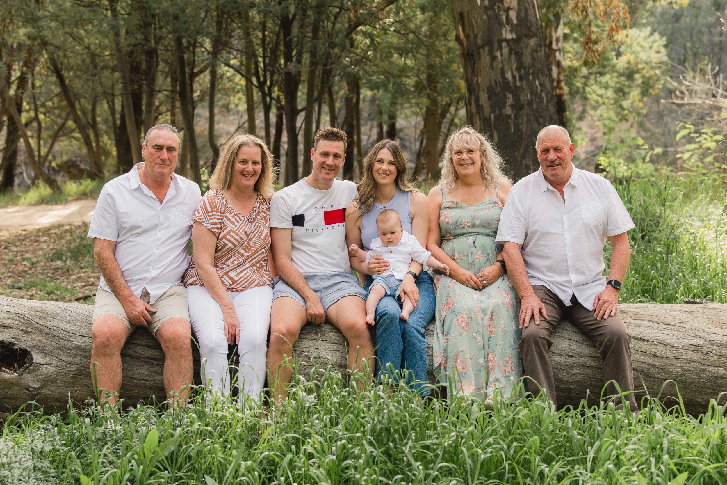 Family of seven sitting on a fallen tree in a wooded park. It includes three men, three women, and a baby, all smiling. The background features tall trees and greenery.