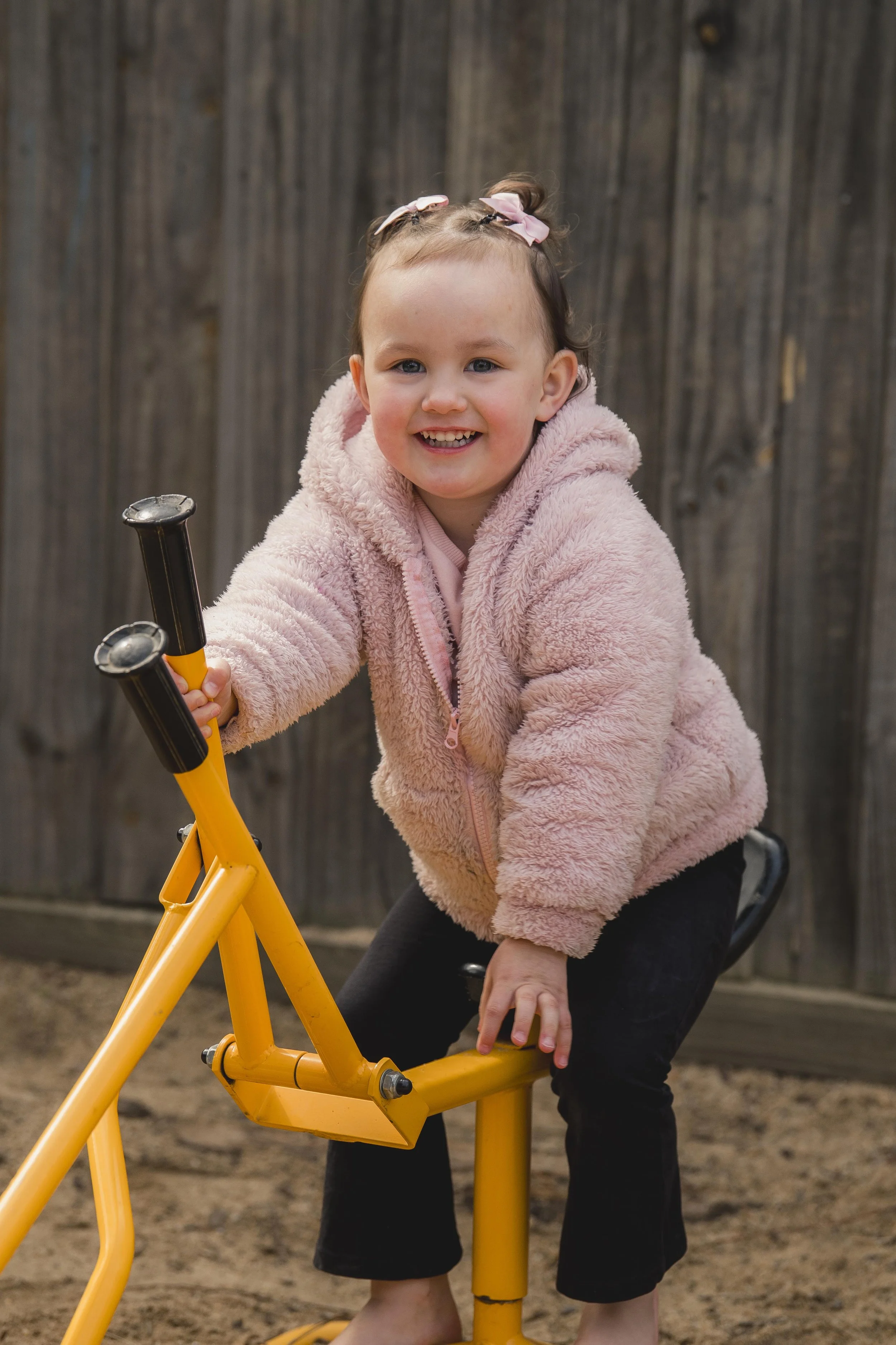 A young girl with a pink curly hair bow riding a yellow pedal-powered ride-on toy in a backyard with a wooden fence.