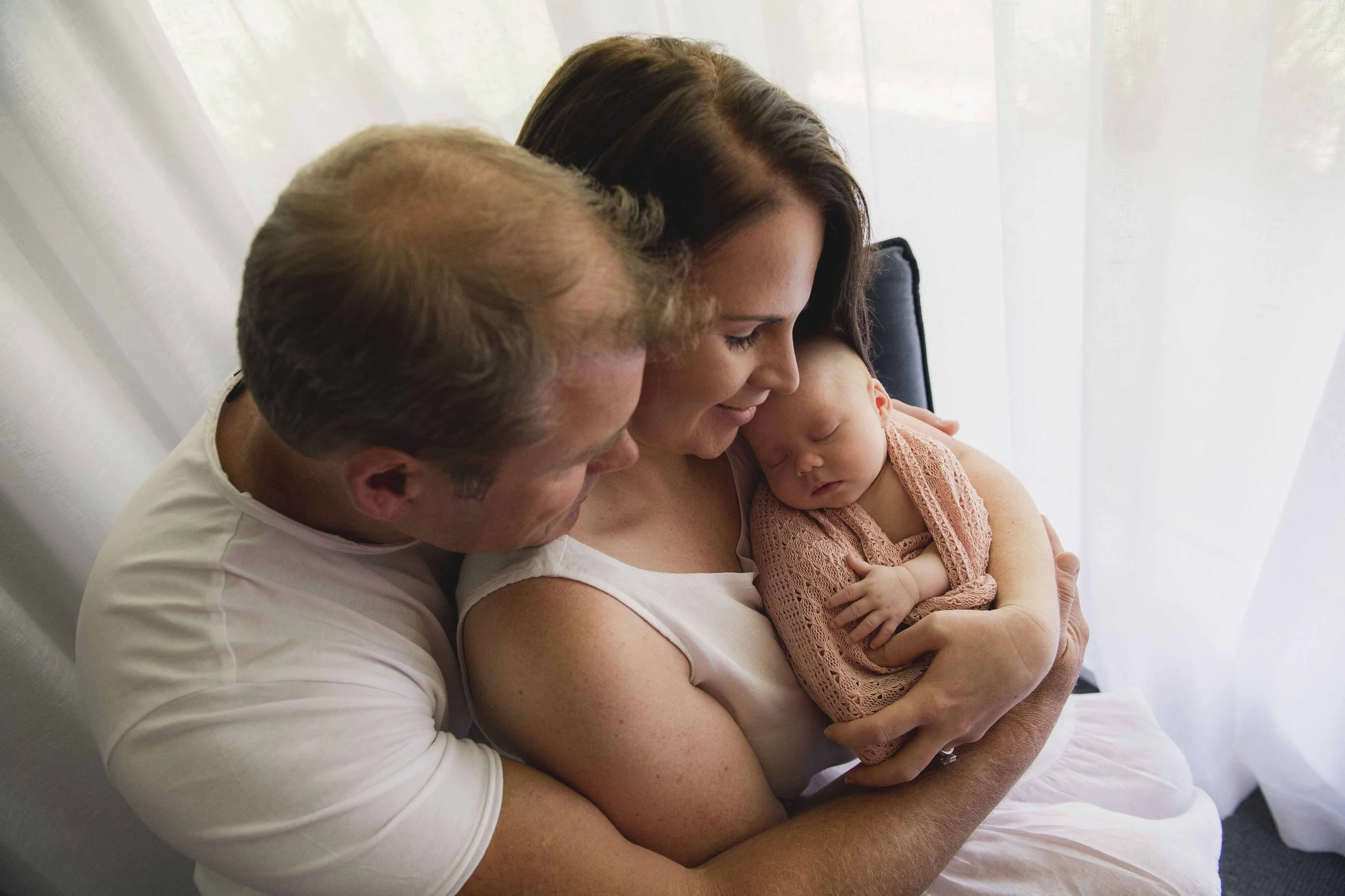 A family of three, a man, a woman, and a baby, sitting close together with their heads touching, sharing a tender moment in front of a white curtain.