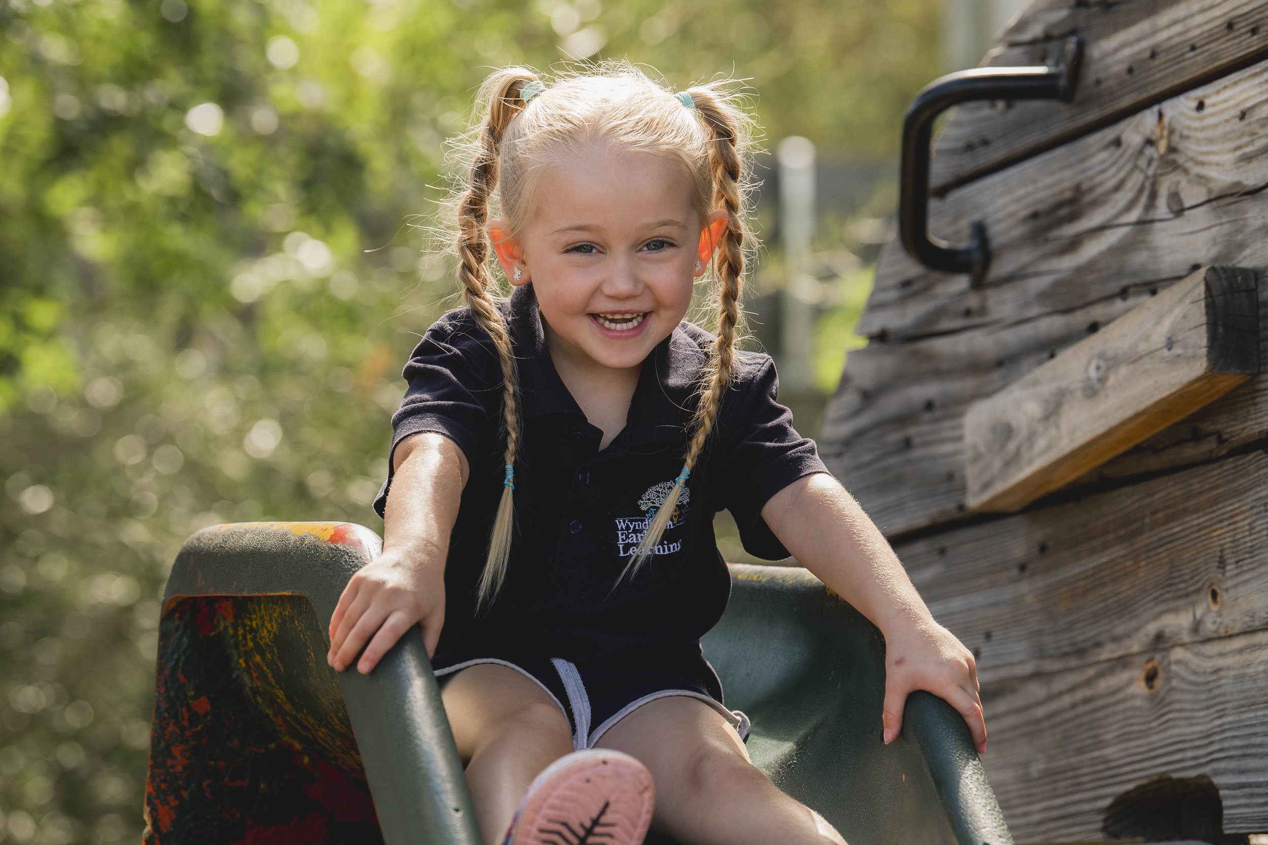 A young girl with blonde hair in braids, smiling, wearing a black T-shirt with a logo, riding a slide outdoors near a wooden structure with a sunny, green background.