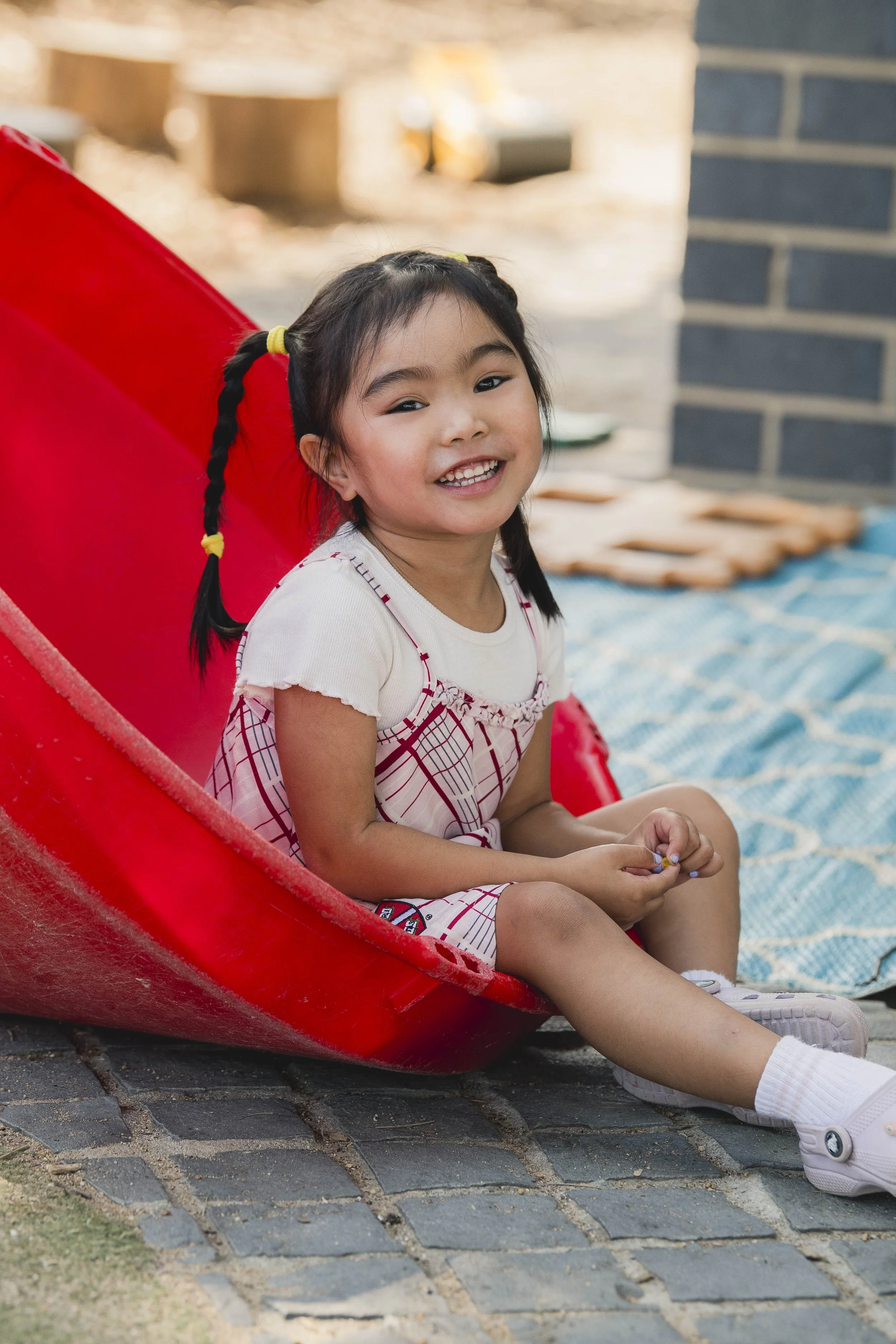 A young girl with braided hair and a white dress with a plaid pattern, sitting on a red slide, smiling happily in an outdoor setting. The background includes a brick wall and a blanket on the ground.