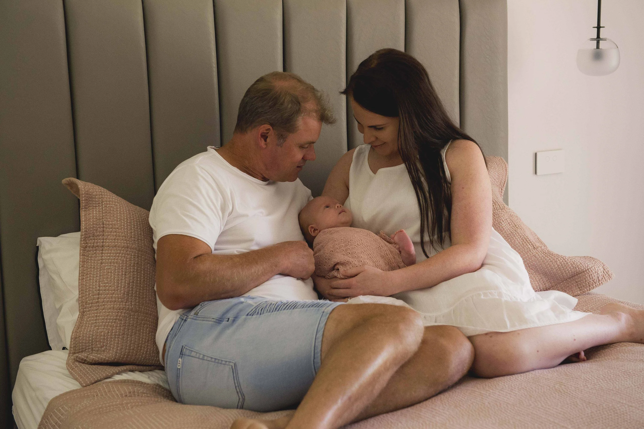 A man and woman sit in bed holding a newborn baby, smiling and looking at the baby. The woman is sitting upright with her dark hair down, and the man is leaning in with his head close to the baby's. The bed has pink pillows and a taupe headboard, wit
