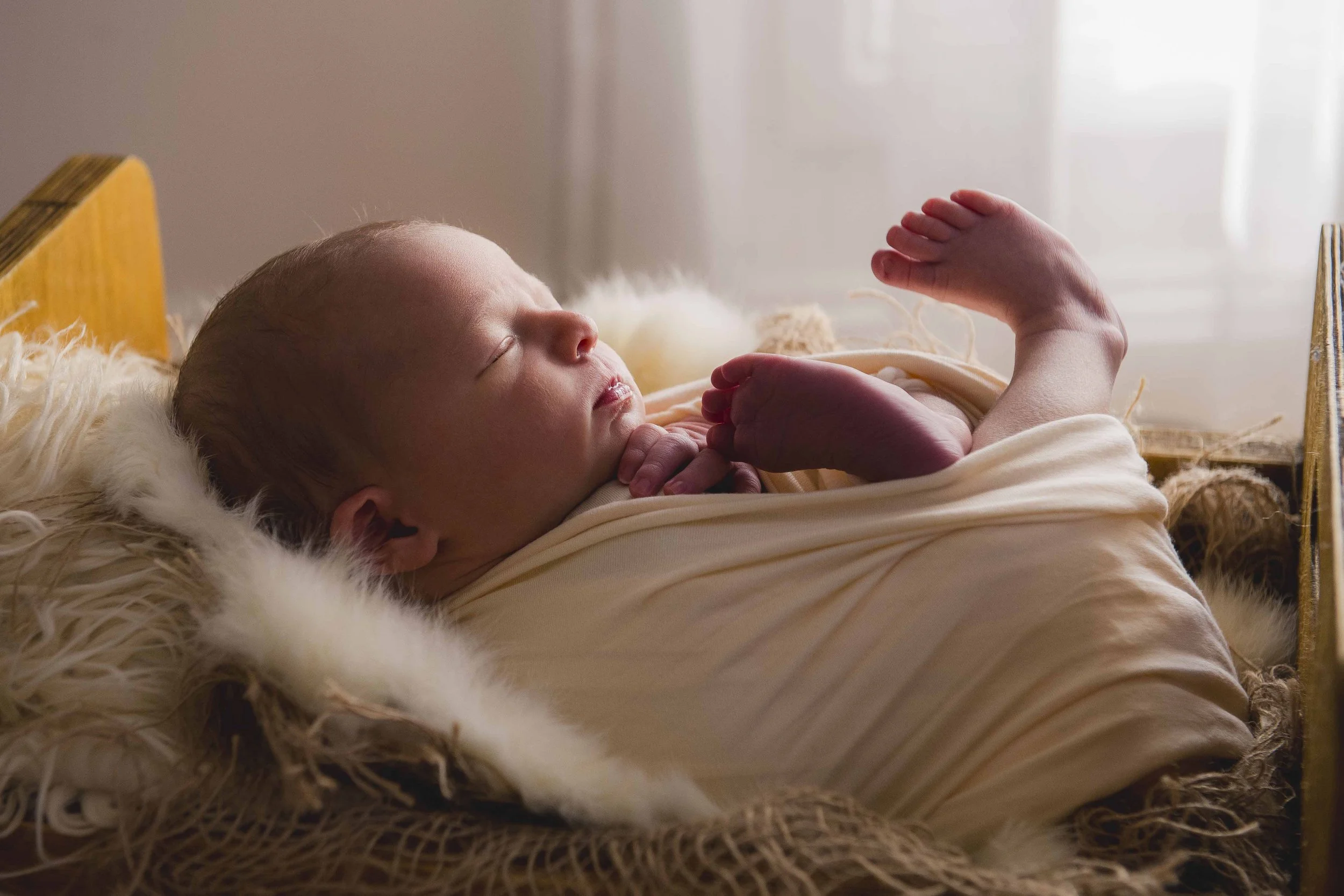 A newborn baby sleeping on a soft, furry blanket in a crib with a wooden frame, with sunlight coming through curtains in the background.