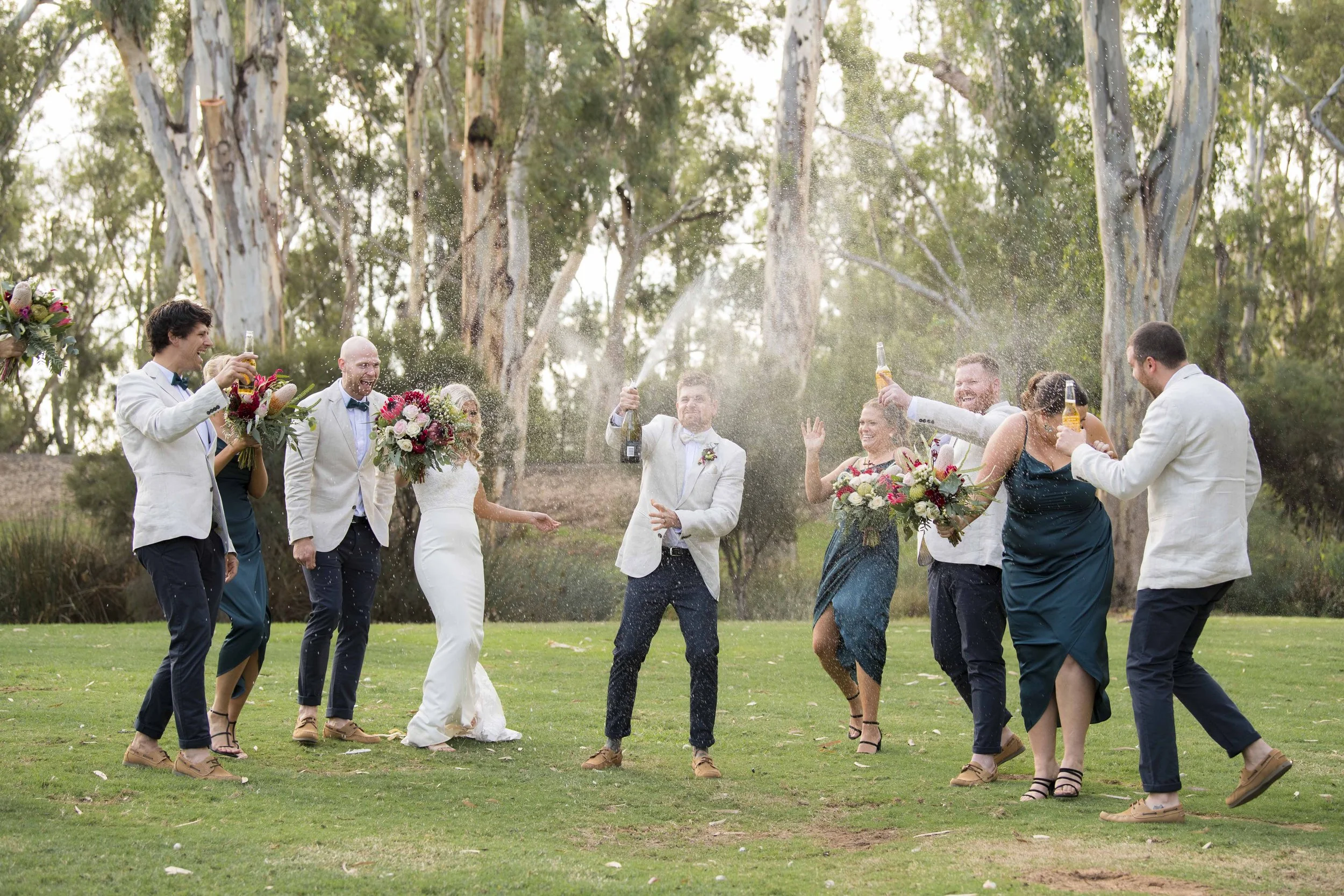 A group of wedding guests celebrating outdoors on a lawn, throwing champagne and smiling.