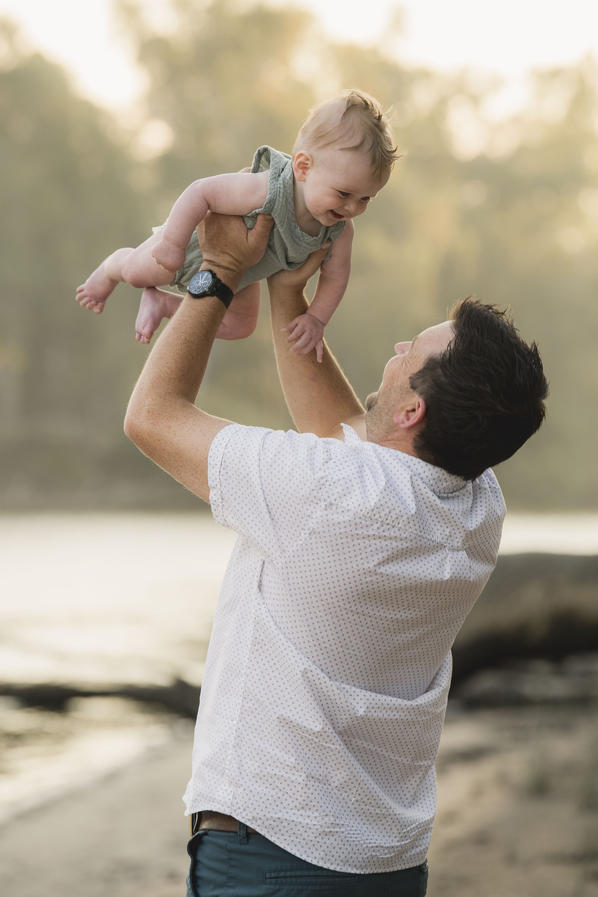 A man lifting a smiling toddler boy in the air outdoors near a body of water during sunset.