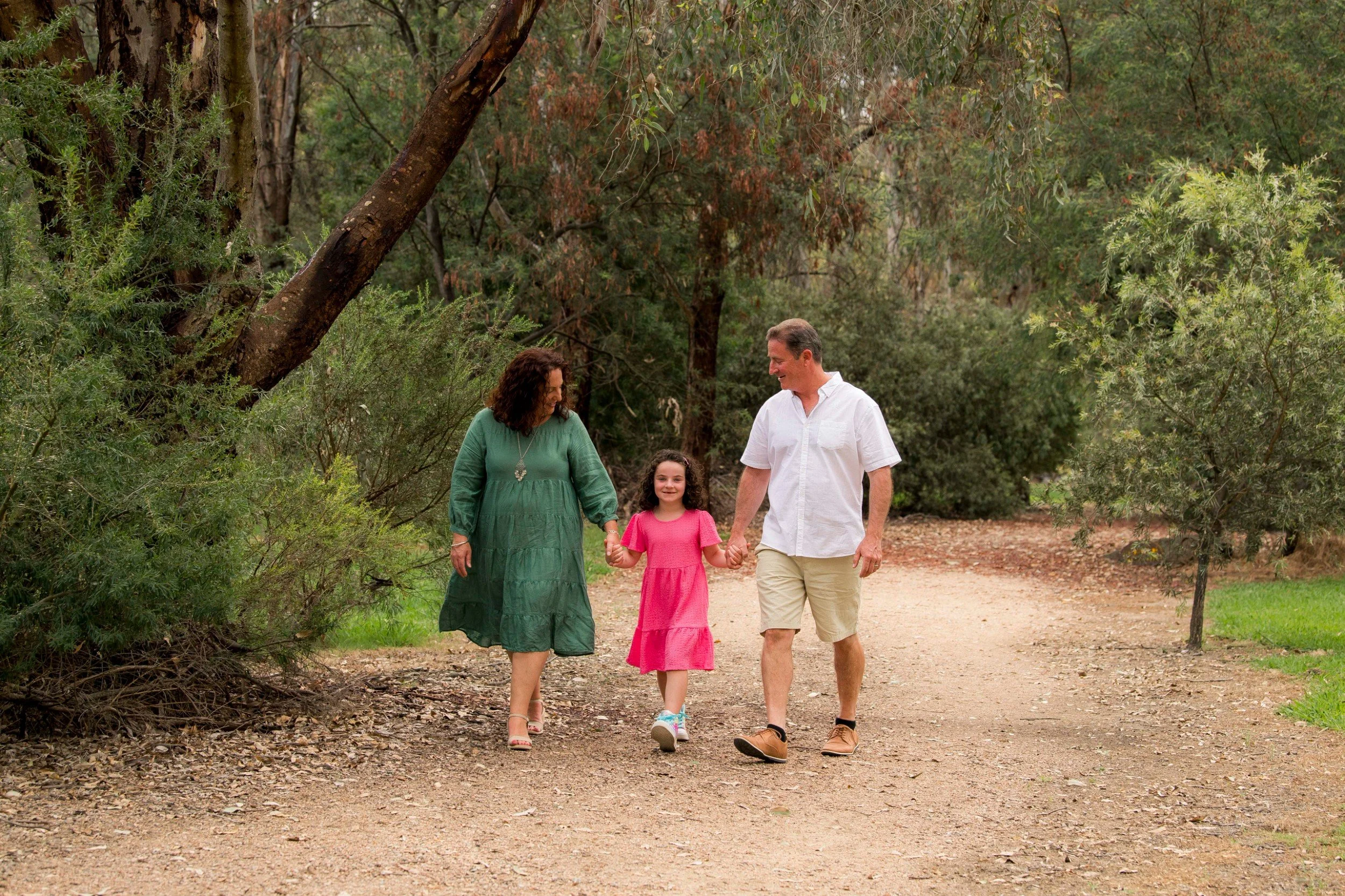 A family of three walking hand in hand on a dirt trail through a wooded area, smiling and enjoying their outdoor walk.