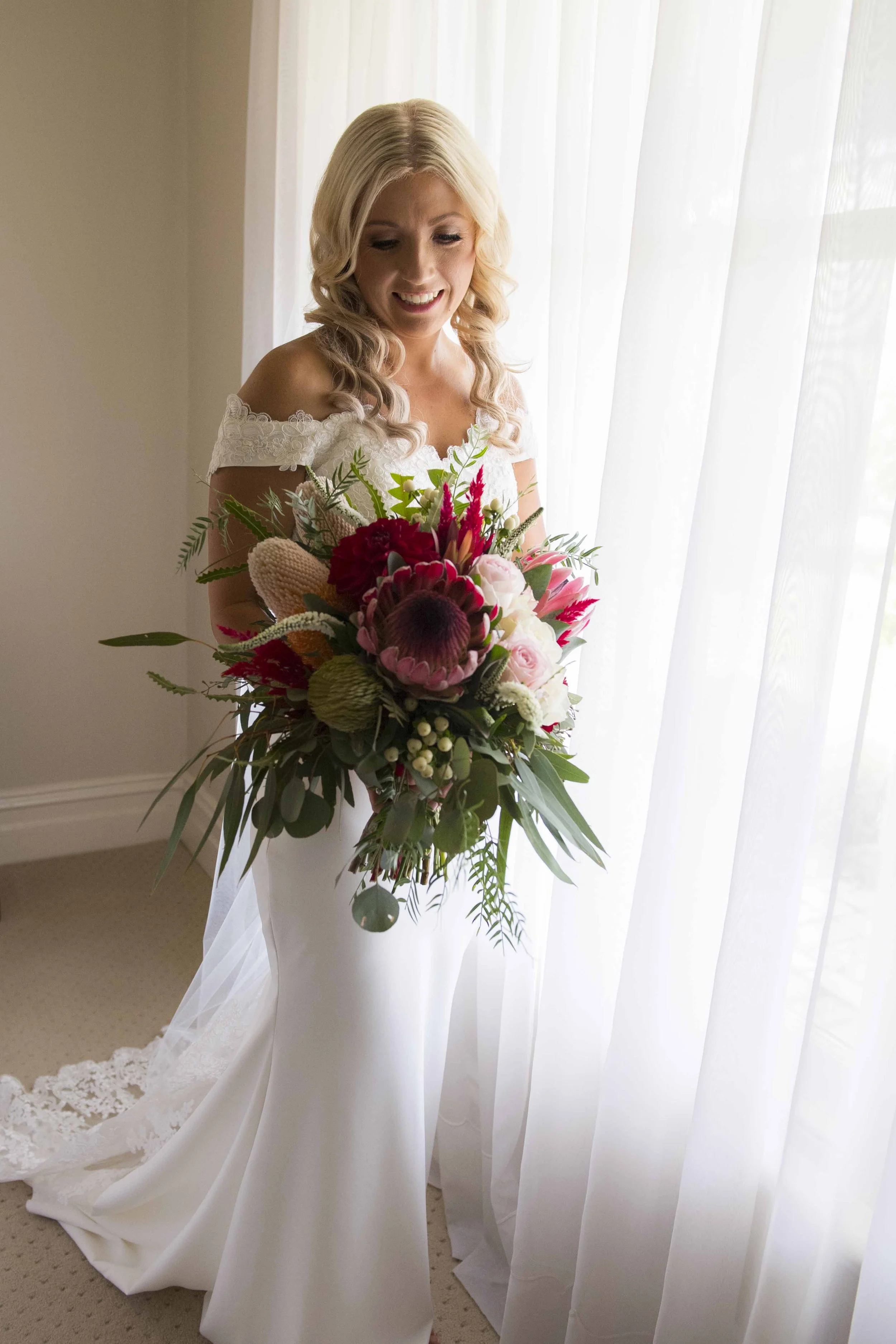 A woman with blonde hair in a white wedding gown holding a large bouquet of colorful flowers in front of sheer white curtains.