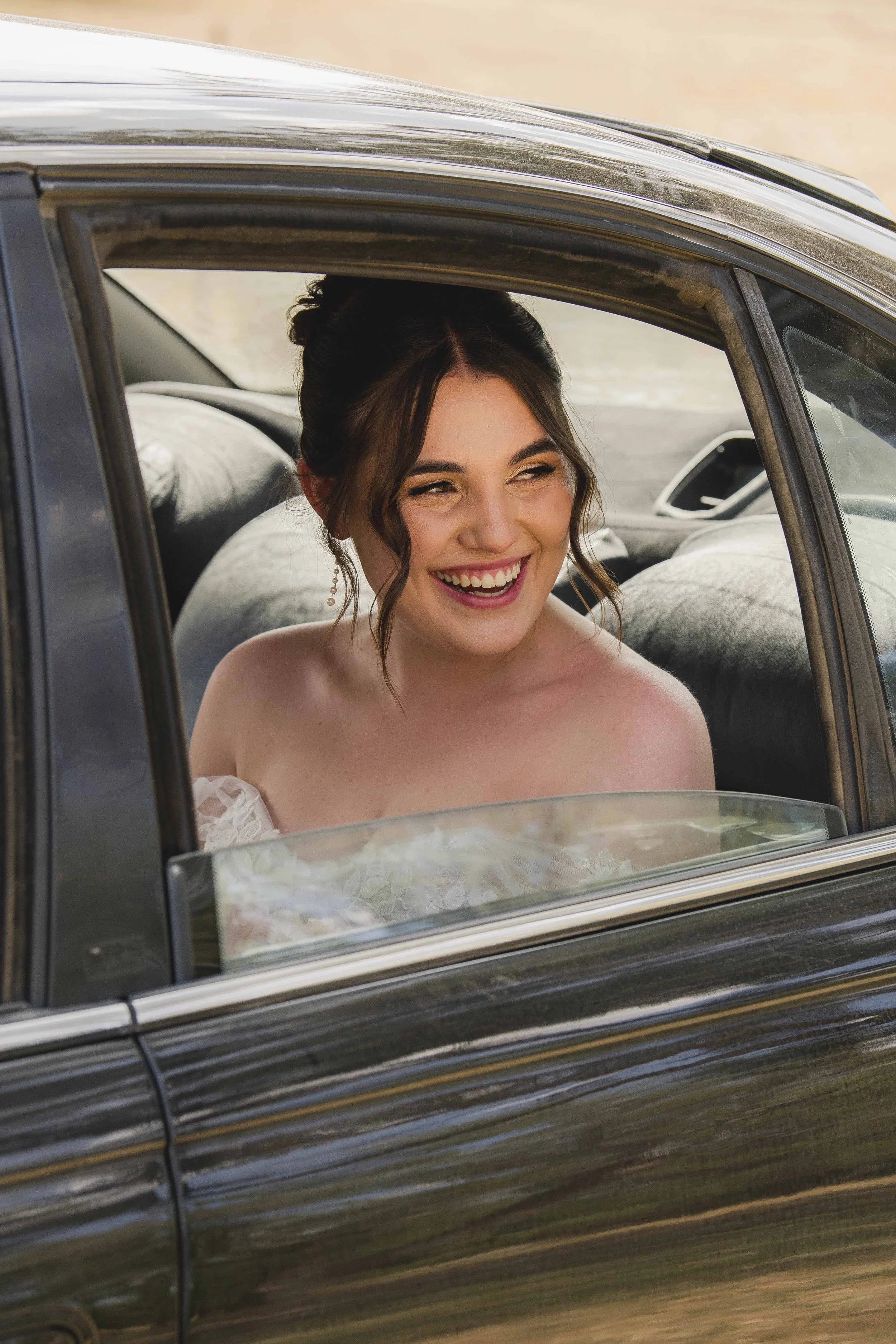 A woman with brown hair styled in loose curls, smiling and winking, sitting inside a classic black car with the window down.