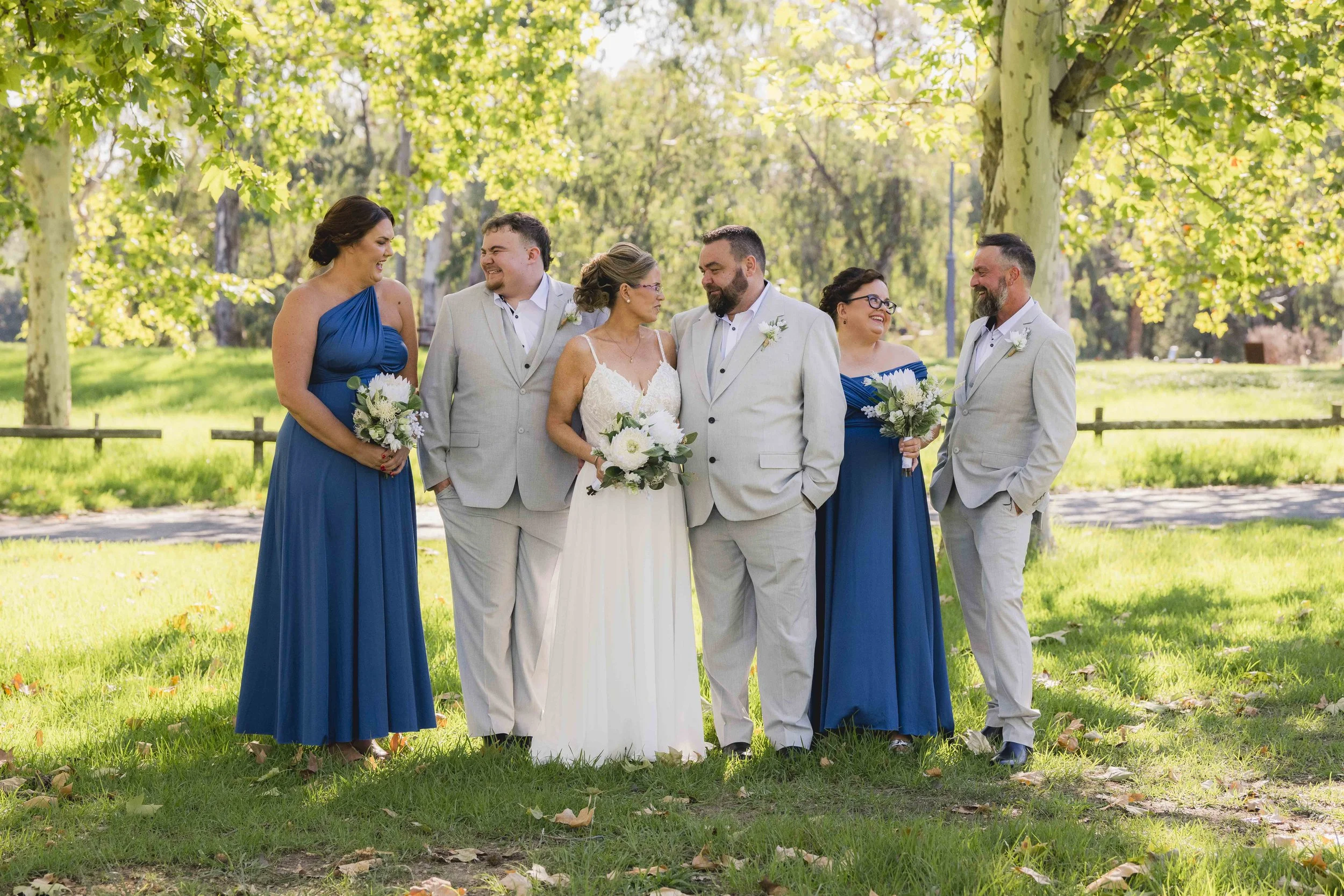 A wedding party standing outdoors under trees, with the bride in a white dress holding a bouquet, surrounded by bridesmaids in blue dresses and groomsmen in light gray suits, all smiling and looking at each other.