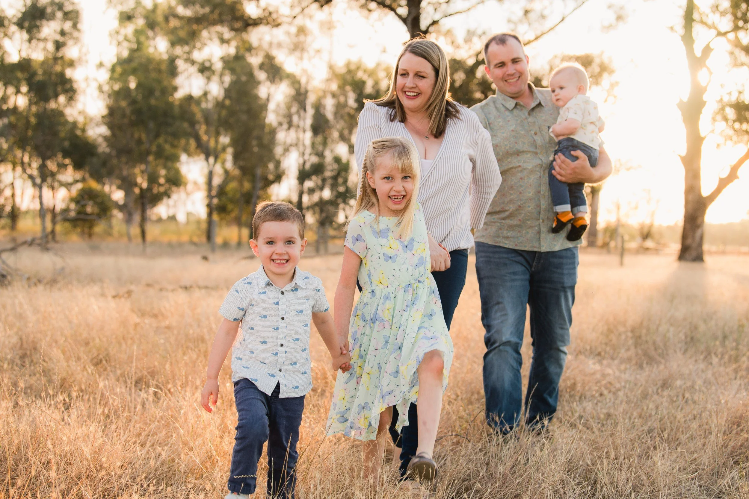 A family of six, including two young children, a woman, a man, and an infant, walking through a field of tall grass during sunset, all smiling and holding hands.
