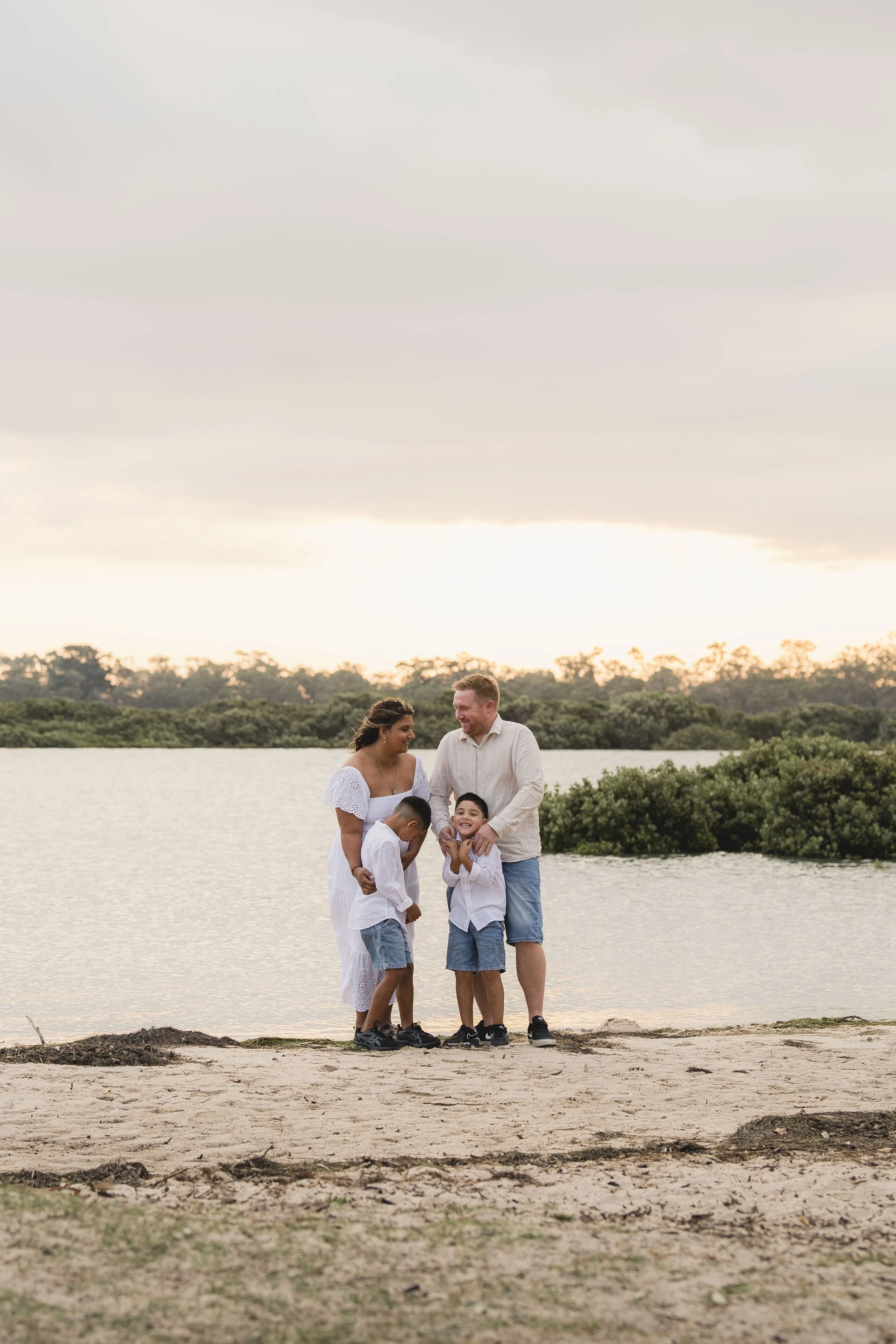 A family of five, including two adults and three children, standing on a beach near a body of water during sunset, engaging with each other and smiling.